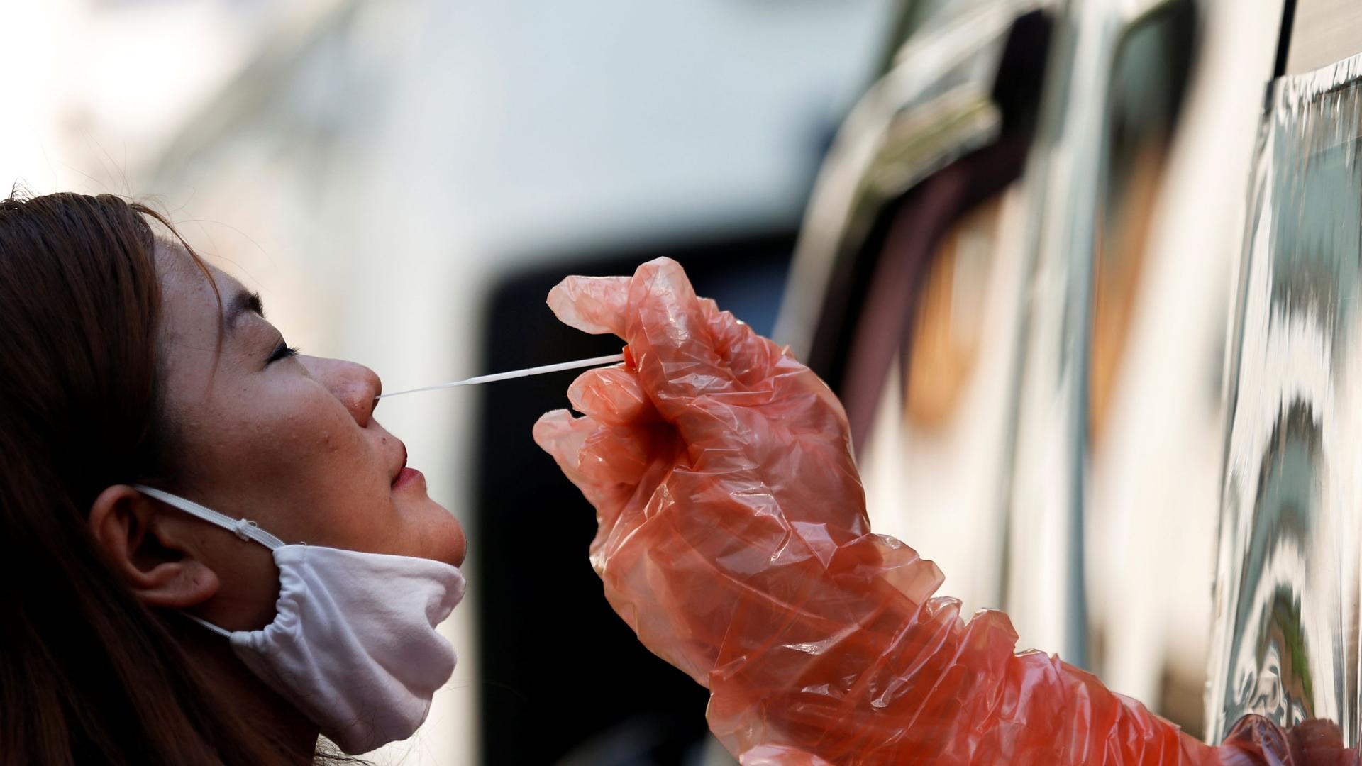 A woman is shown leaning her head back slightly with a protective mask pulled down and a health care worker reaches out with a swab.