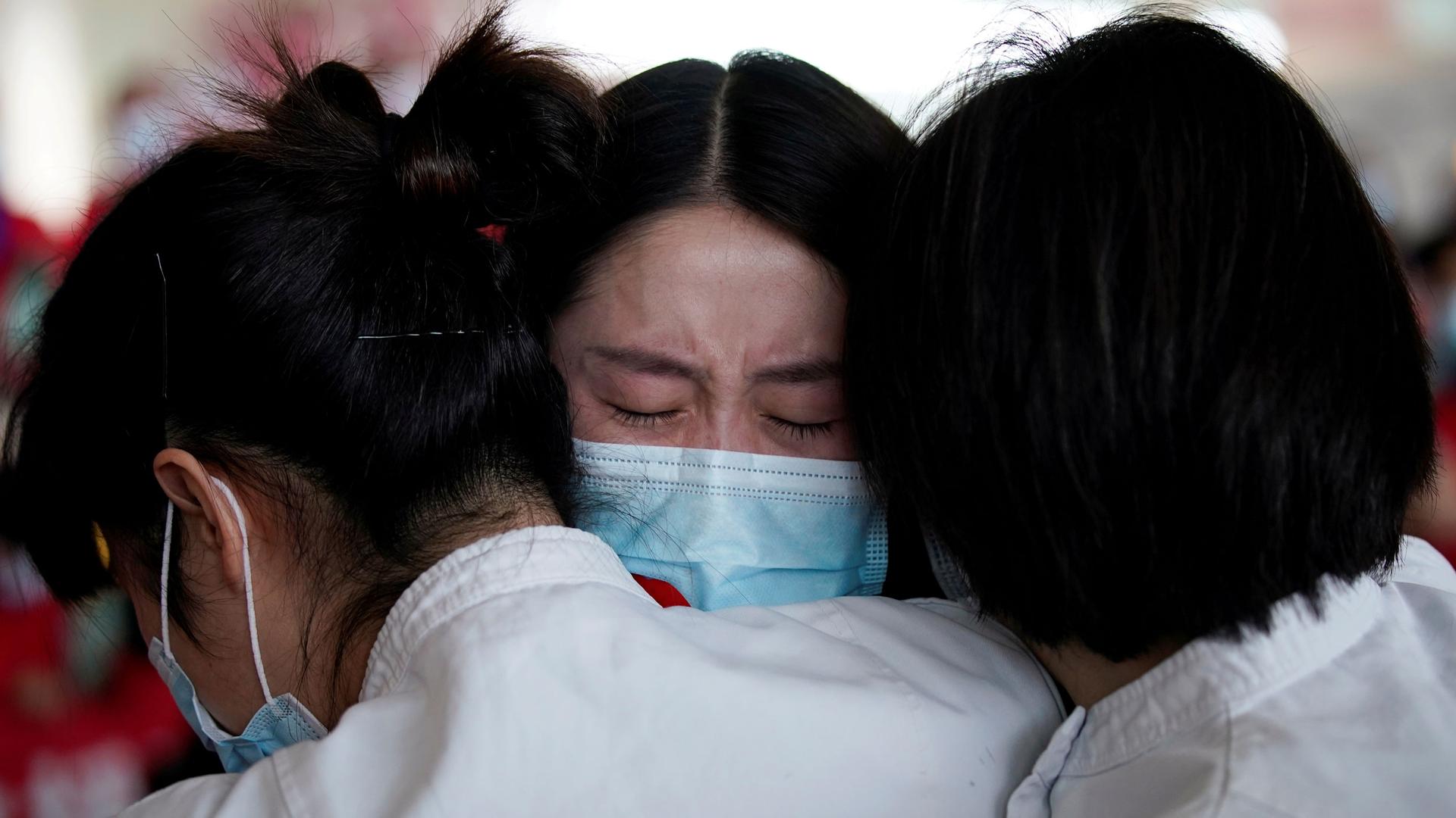 A close-up photograph of three women are shown hugging and wearing protective face masks.
