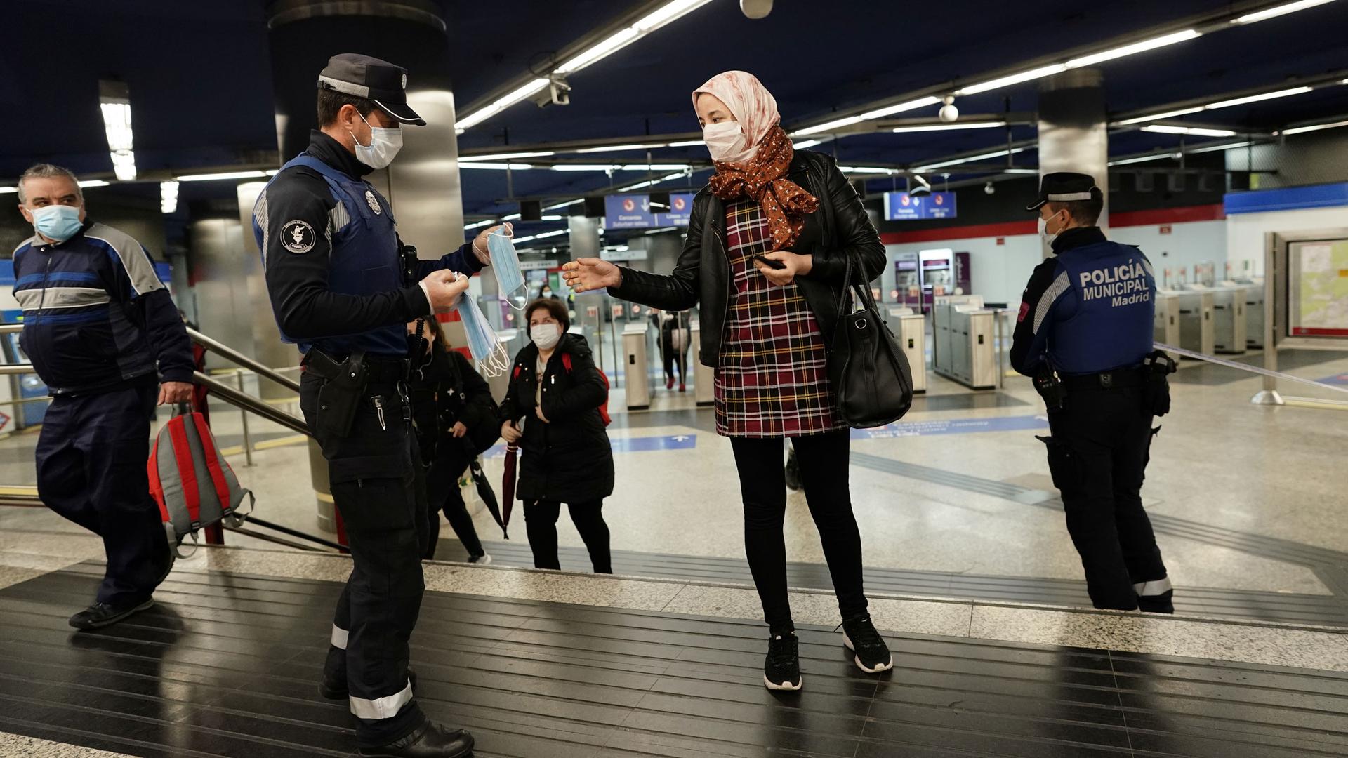 A police officer is shown holding several protective face masks and handing one out to a woman standing adjacent.