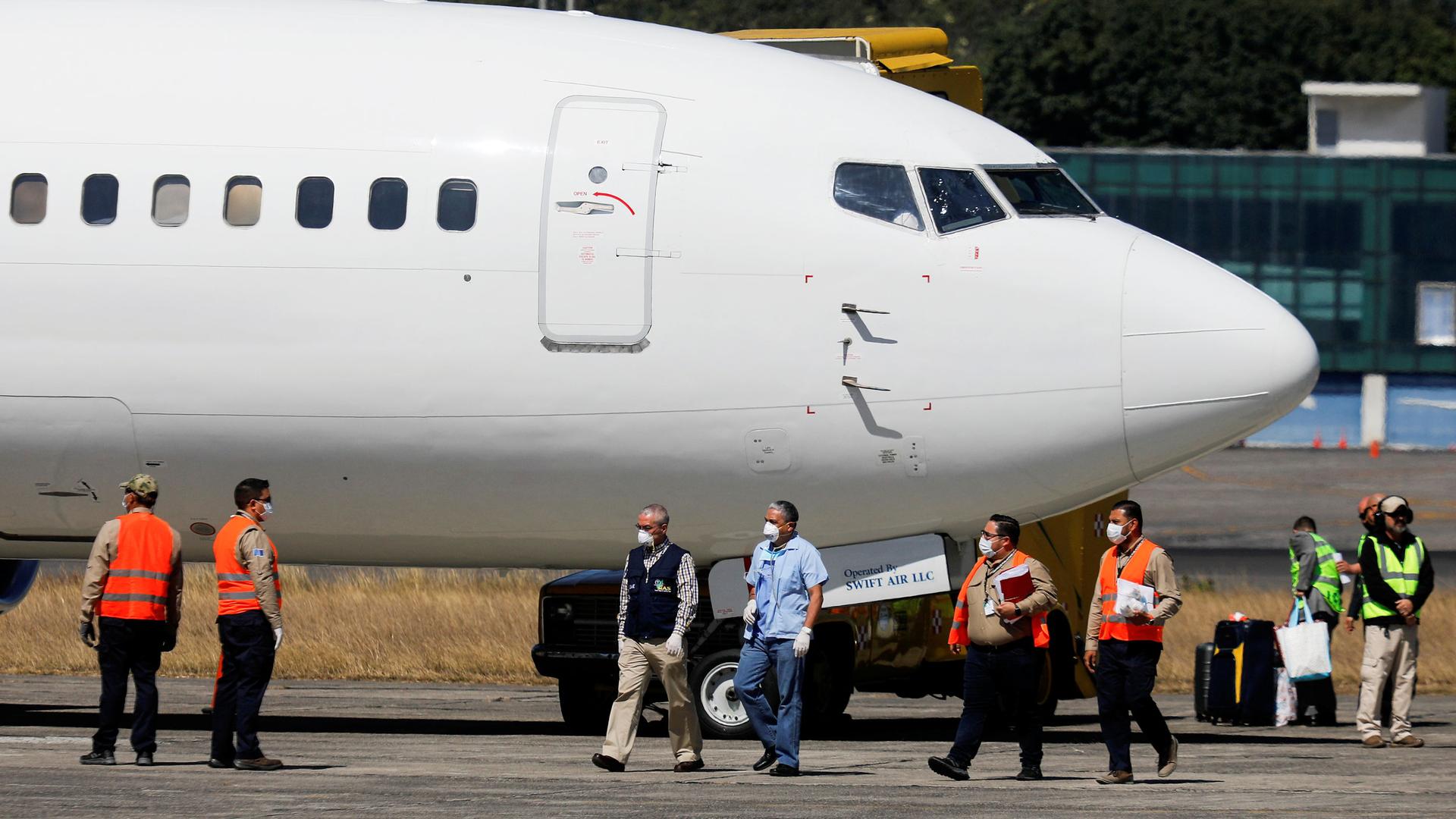 Government officials, wearing protective masks, stand next to a plane