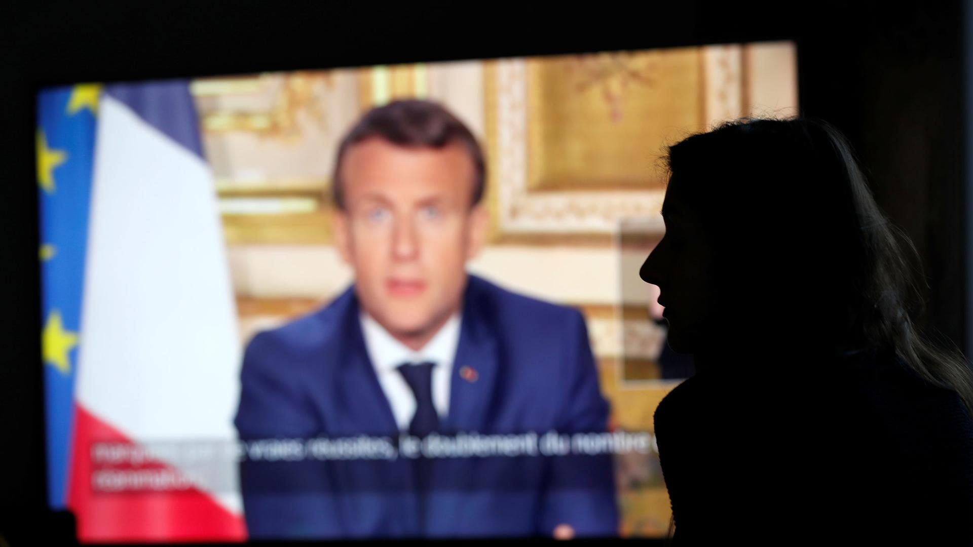 French President Emmanuel Macron is seen on a TV screen in soft focus with a person in shadow in the nearground.