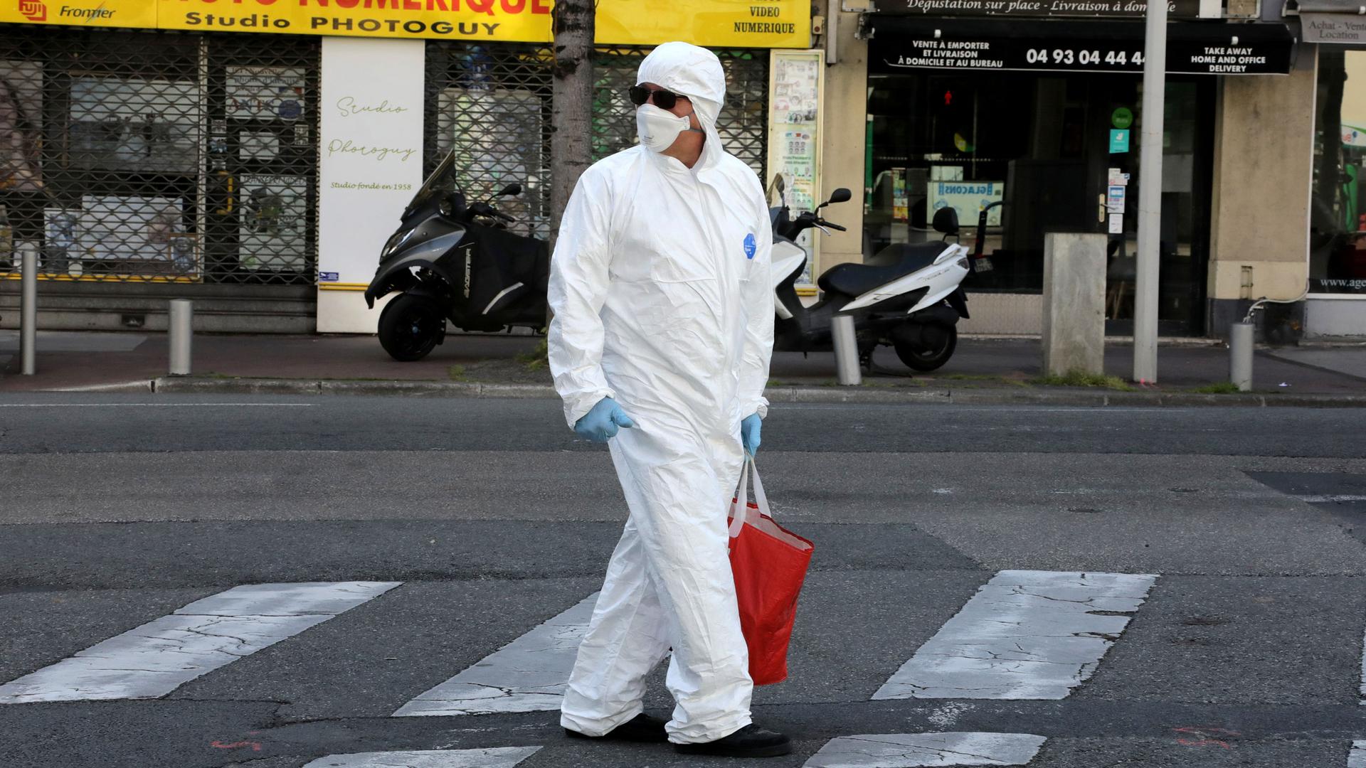 A man wearing a white protective suit and face mask with sunglasses is shown looking over his shoulder while walking in a street.