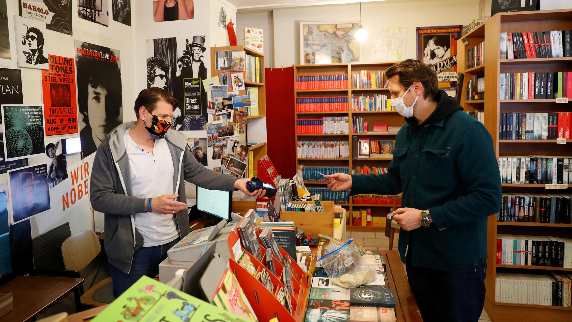 A man holds a card reader as another pays for a purchase in store with books and music posters. 