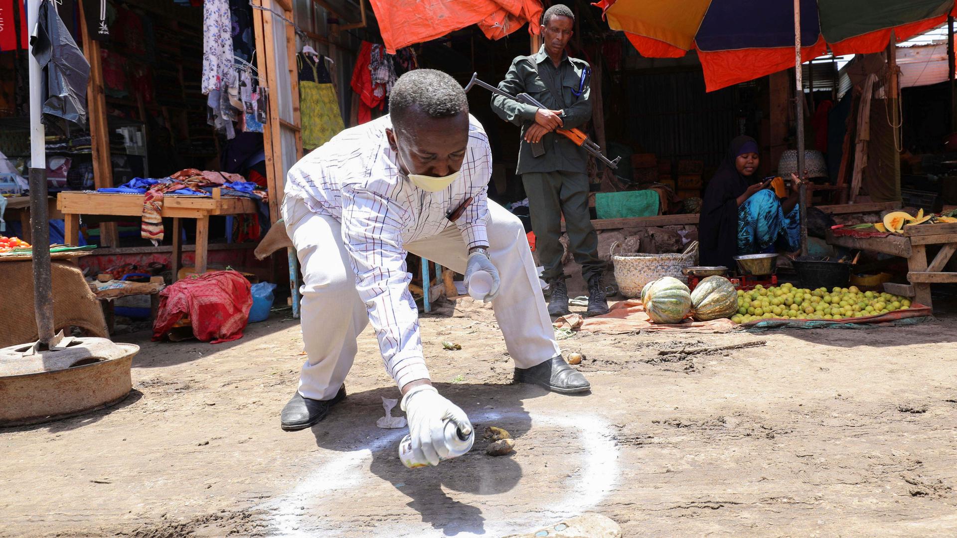 A man is shown bent over and spaying a white circle on the ground with a man nearby holding a weapon.