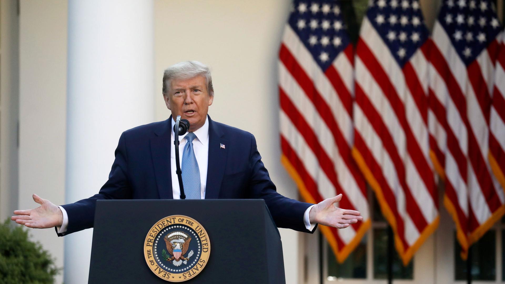 US President Donald Trump is shown standing at a podium with the US Presidency emblem on it with both of his arms outstretched.