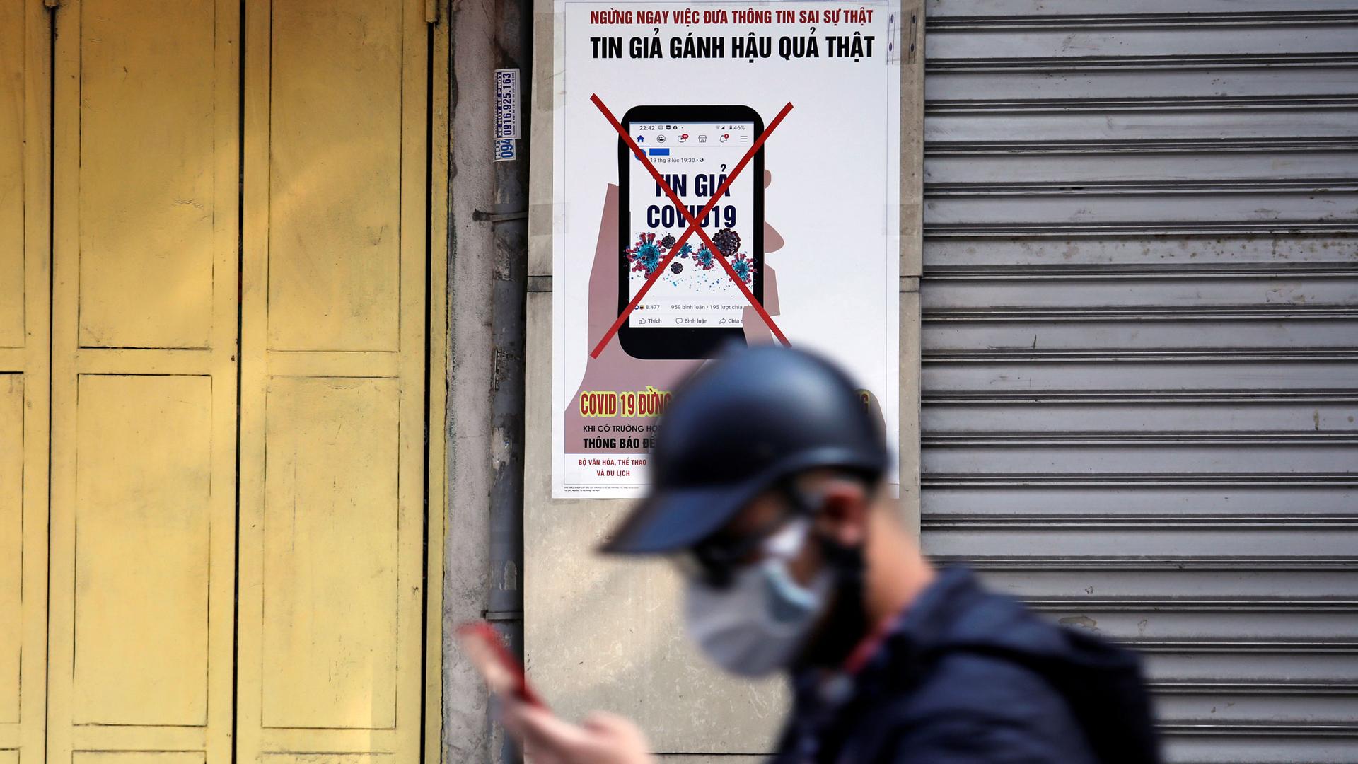 a man with a cellphone and a face mask walks in front of a sign in Vietnamese discouraging the spread of misinformation. 