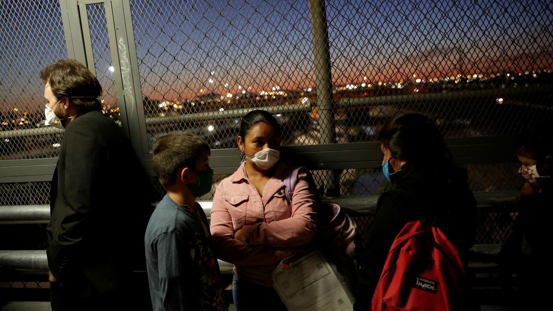 A line of people are shown wearing protective face masks and standing next to a fence.
