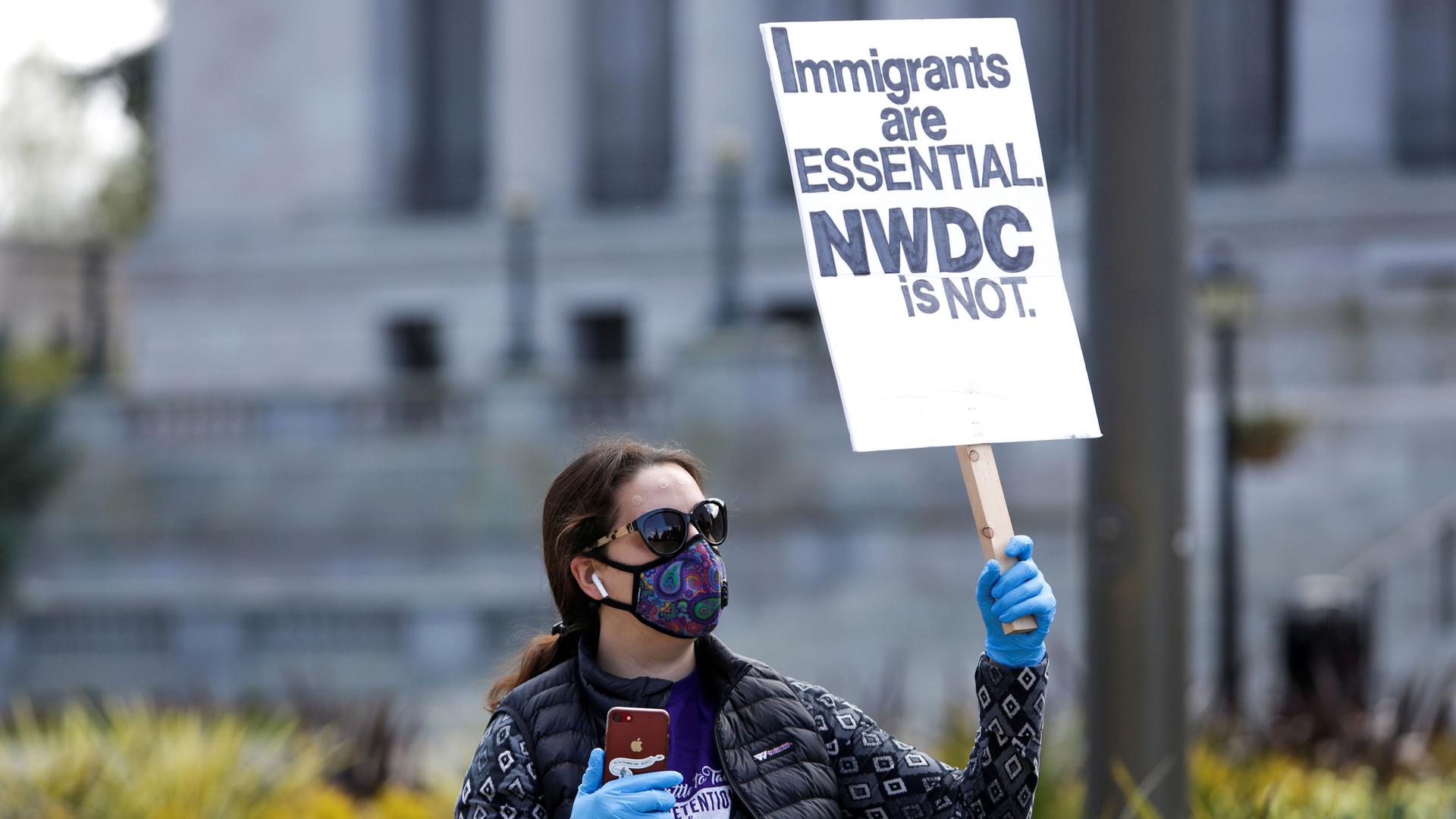 A woman in a mask and gloves holds a sign reading "immigrants are essential NWDC is not"