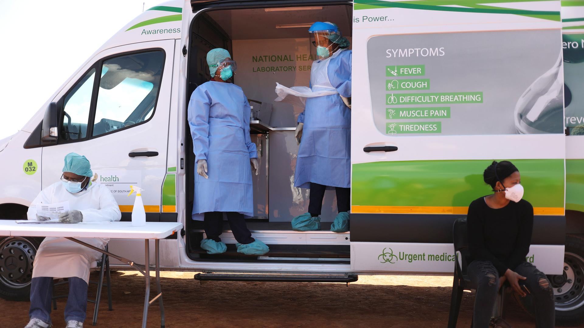 Two people are shown standing the side opening of a van and wearing protective medical gear with two more people sitting outside of the van.