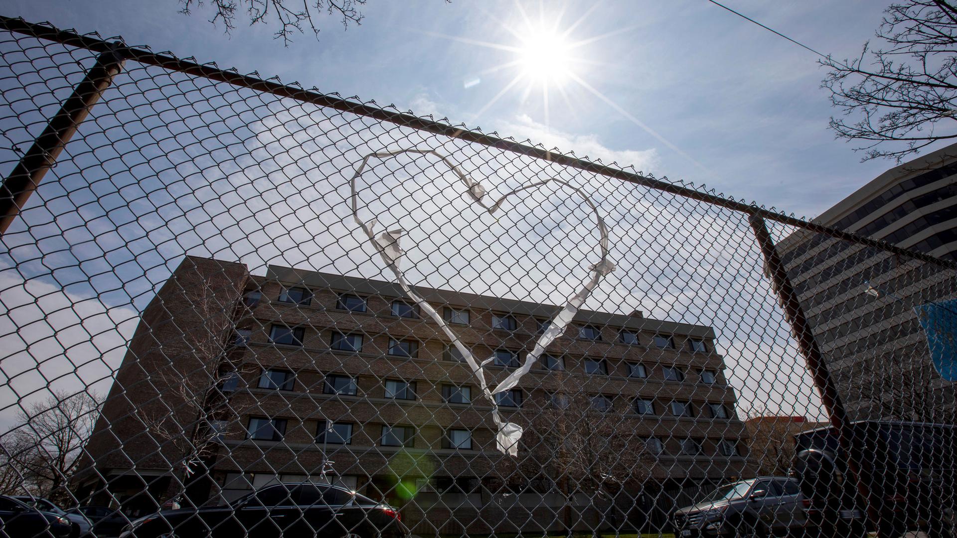 A chain-link fence is shown with a clothe heart woven into it and a brick building in the background.