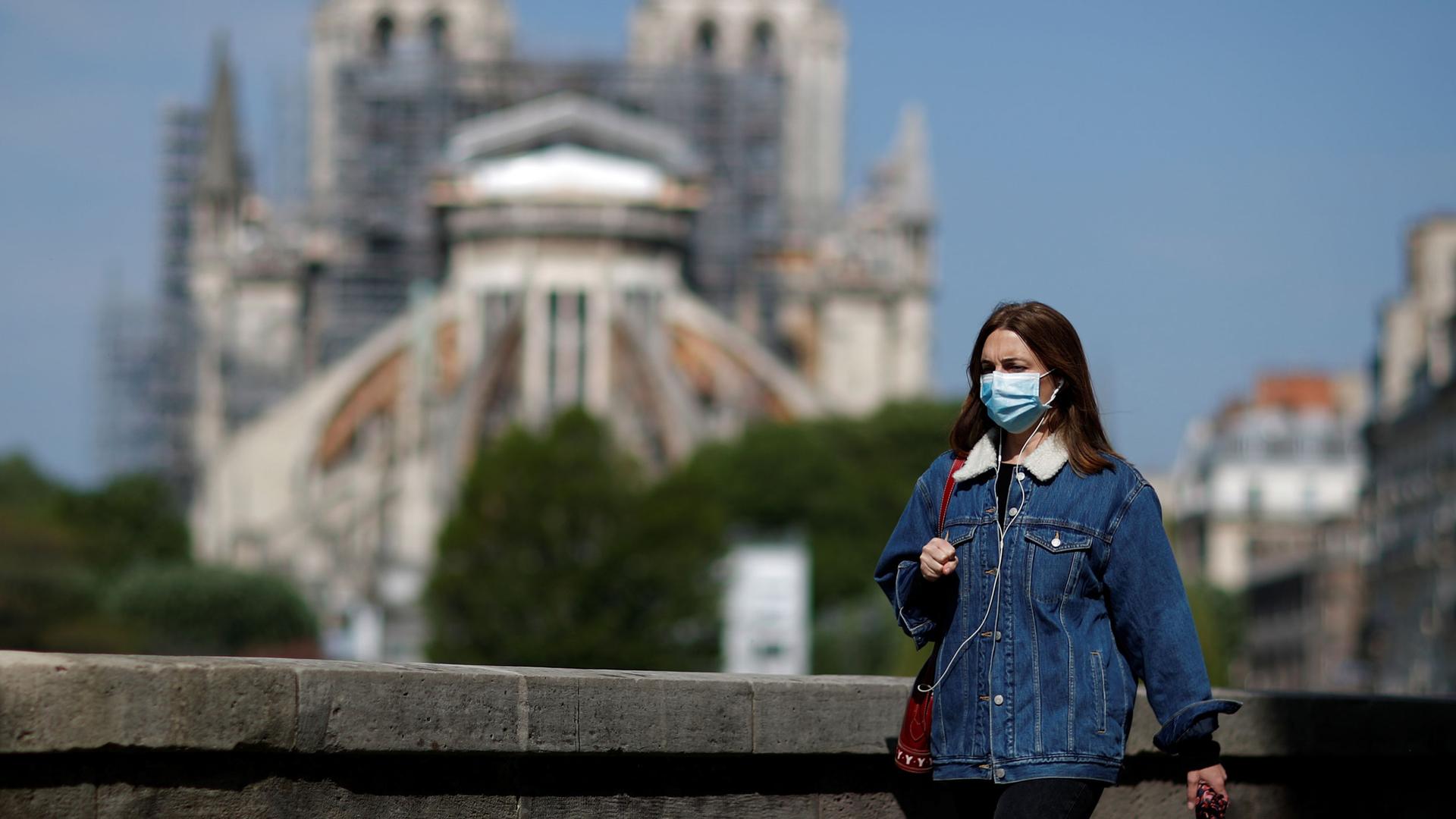 A woman, wearing a protective face mask and a jean jacket is shown walking past Notre-Dame Cathedral in the background in soft focus.