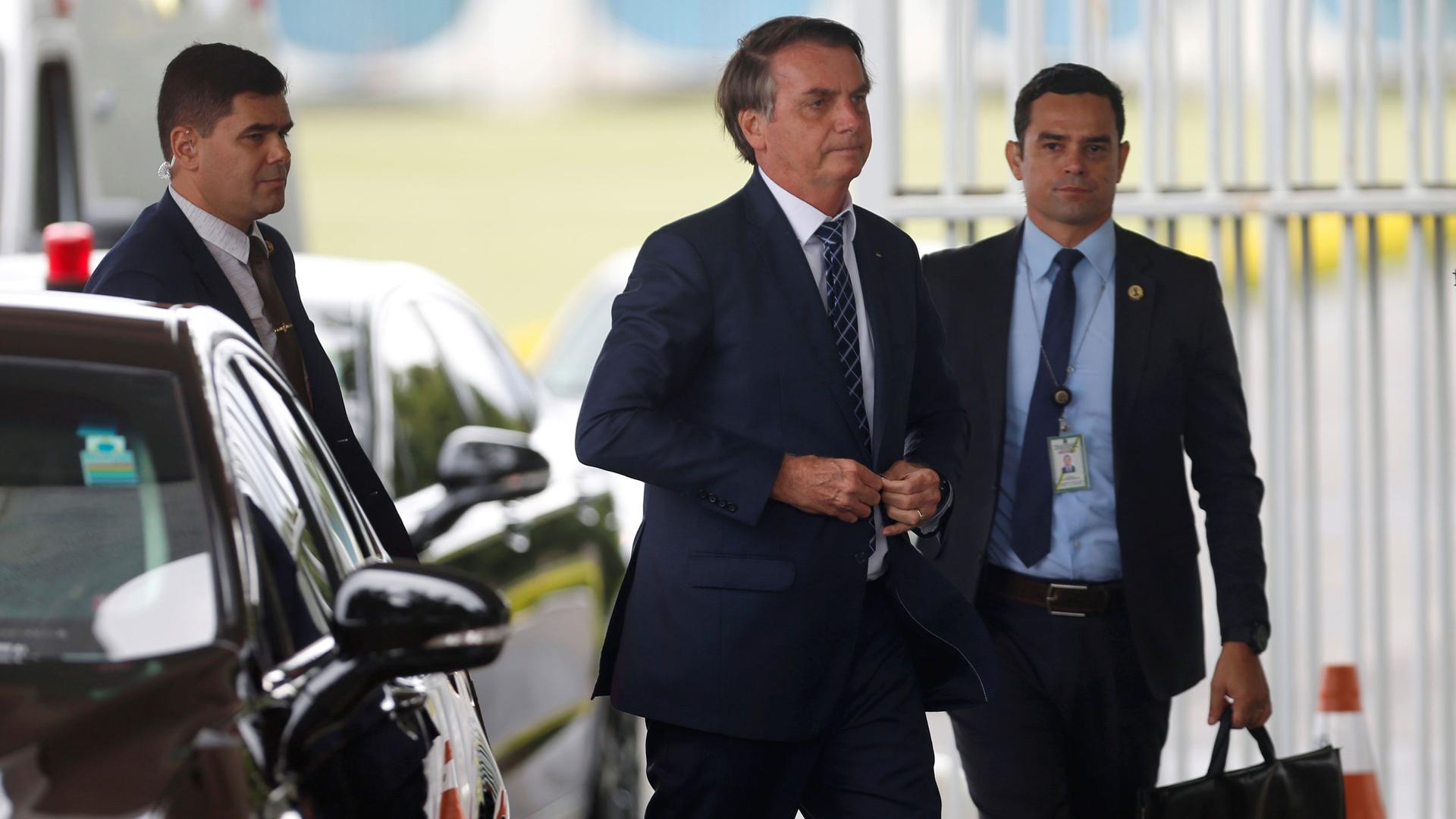 Brazil's President Jair Bolsonaro is shown buttoning his suit jacket and walking with security detail on either side.