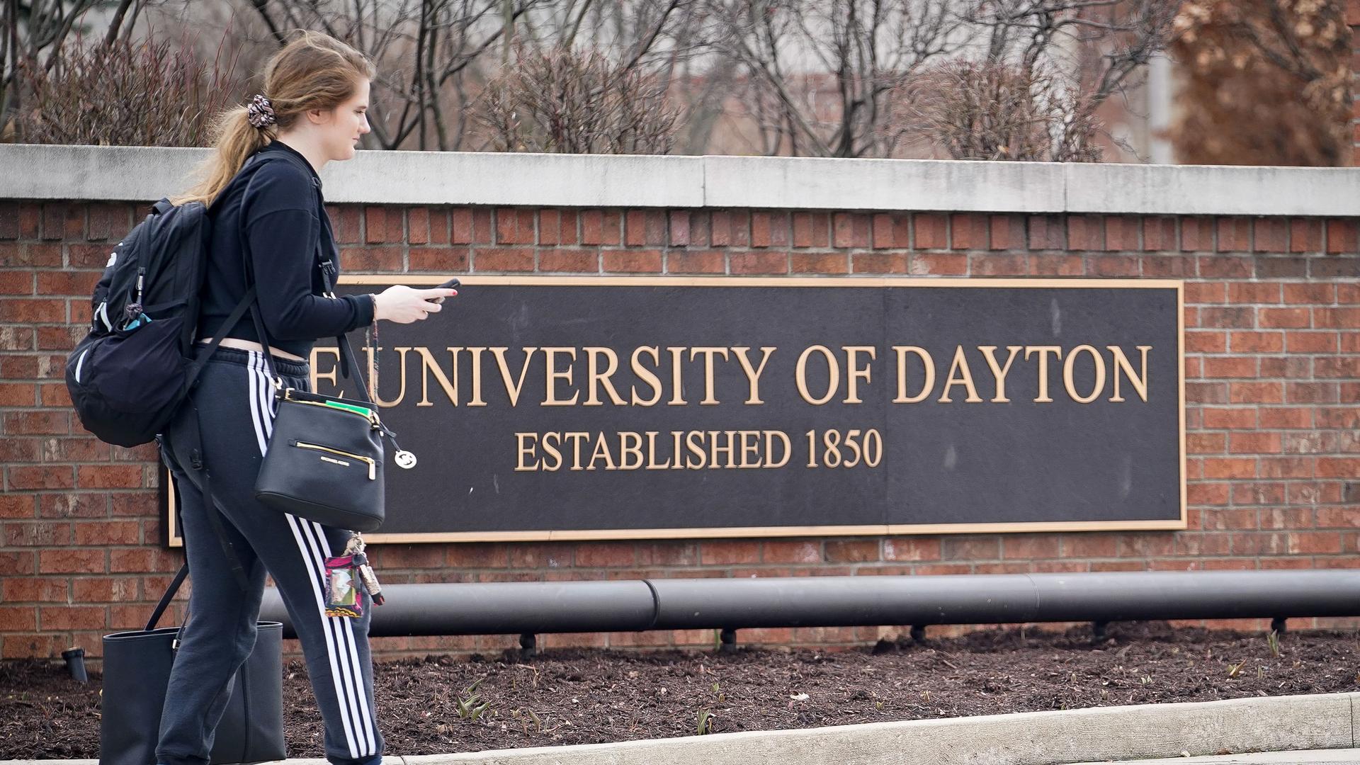 A student carries her bags in front of a sign for the University of Dayton
