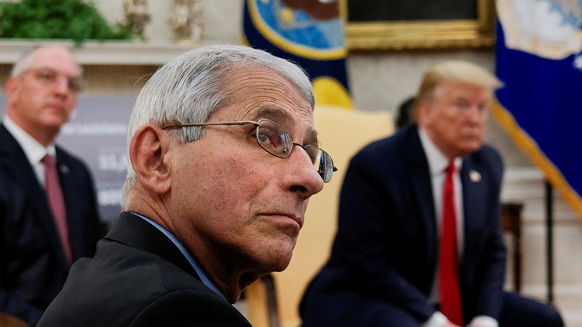 A close up photograph of Dr. Anthony Fauci who is wearing a dark suit and glasses with President Donald Trump shown in soft focus in the background.