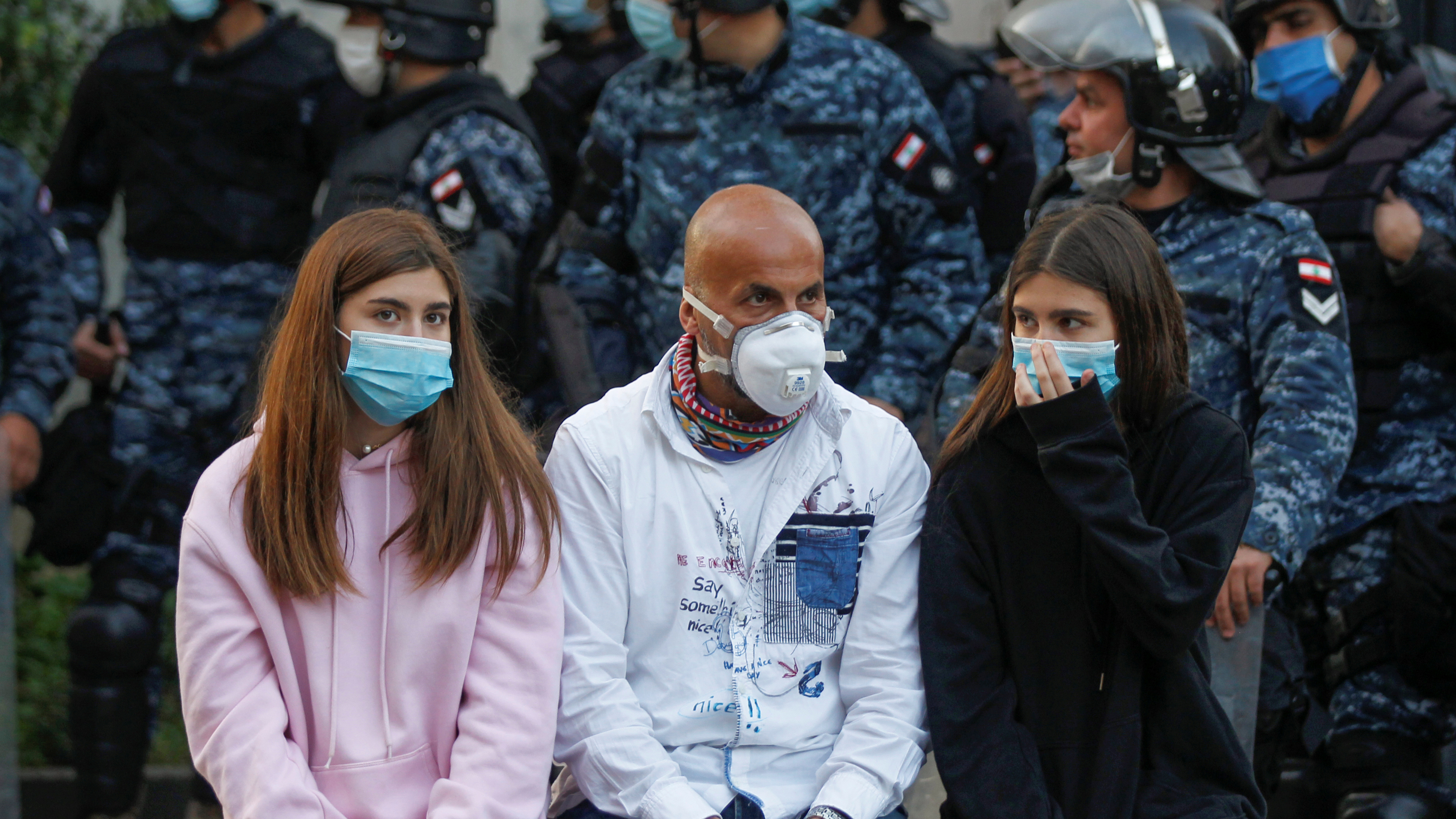 Three people wear face masks with soldiers standing behind them 
