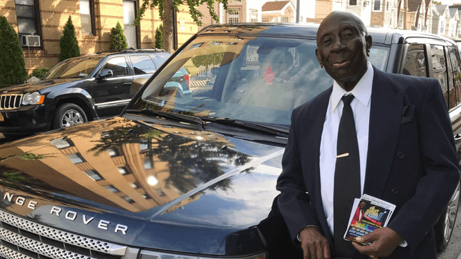 A Black man in a suit stands by a Range Rover holding a book