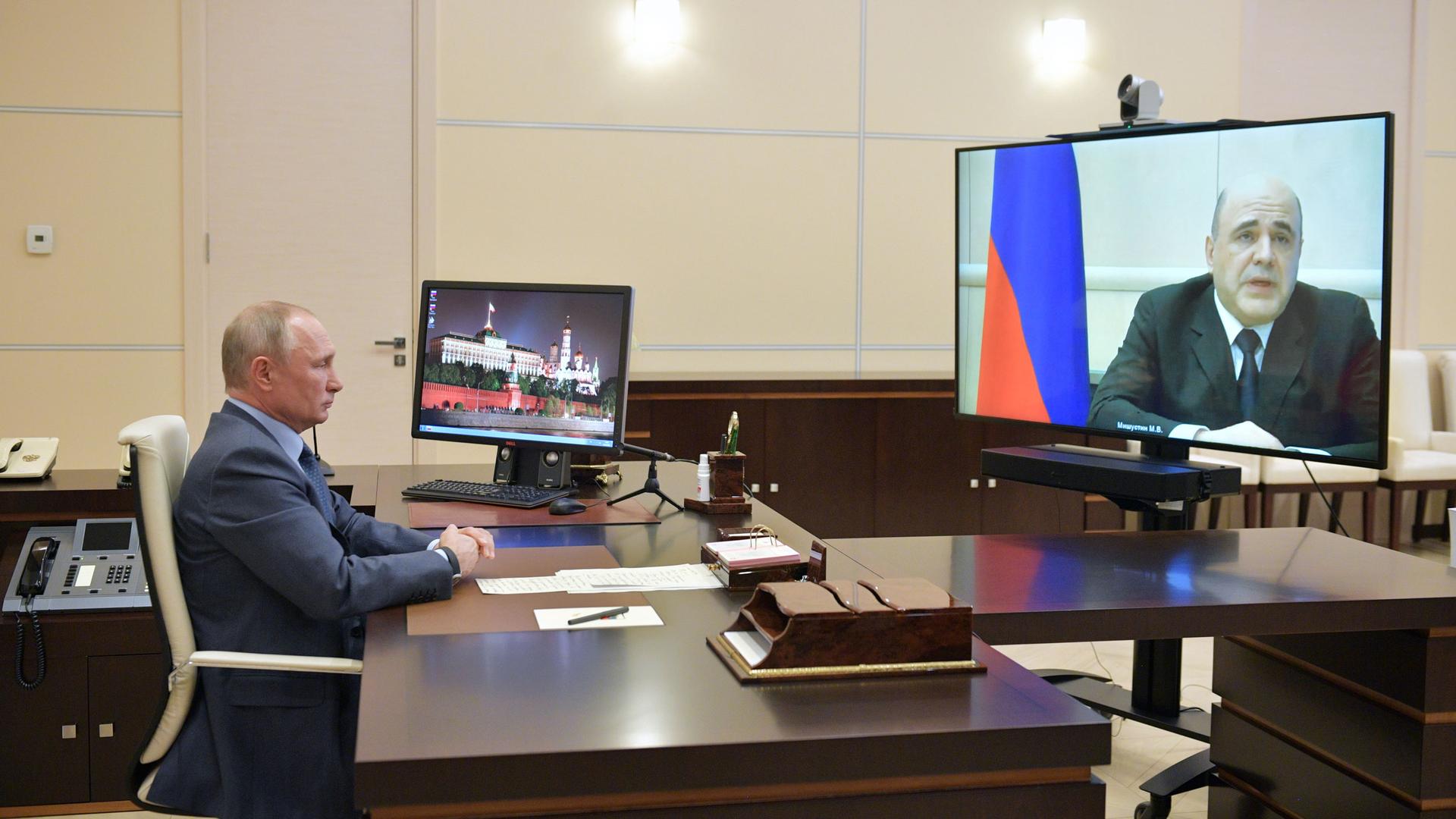A man at a desk looks at a man on large screen 