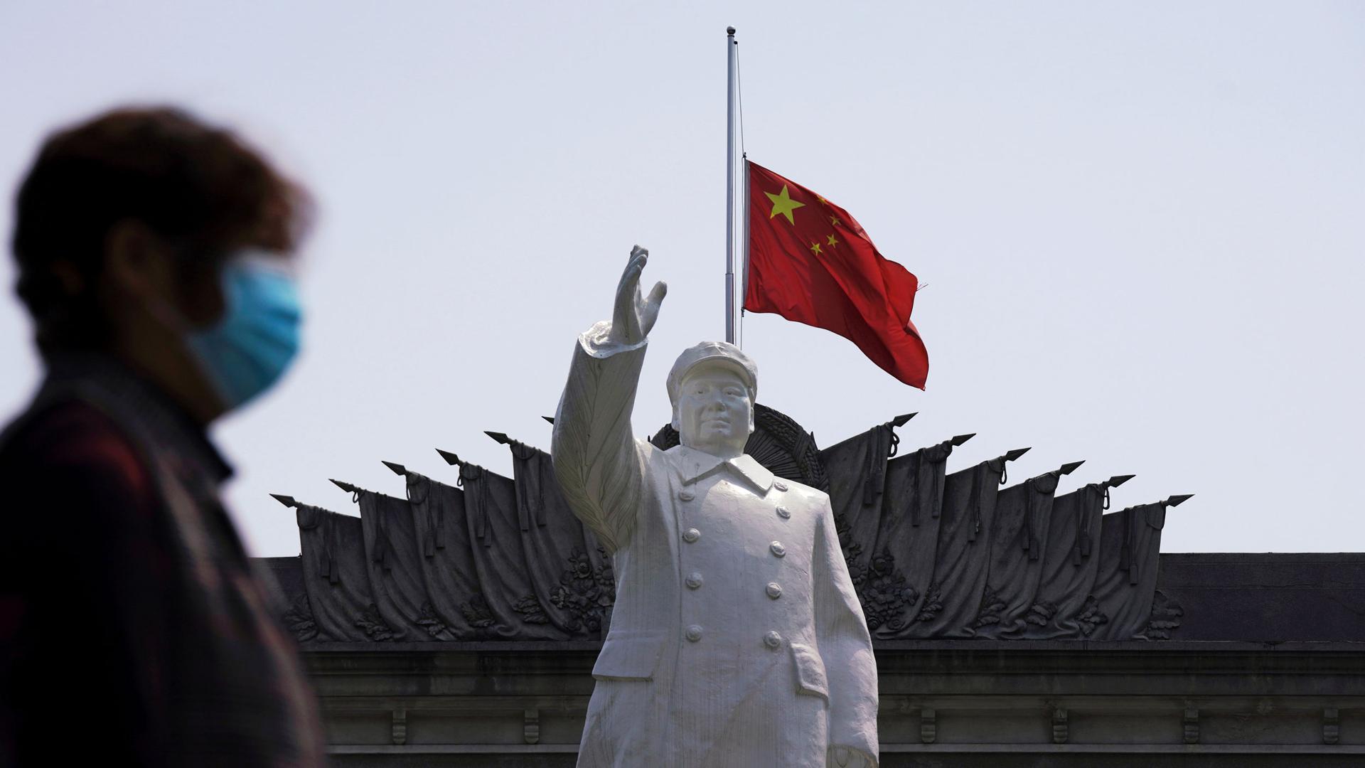 A person wearing a protective face mask is shown in soft focus walking past a statue of late Chinese chairman Mao Zedong with the red Chinese flag flying.