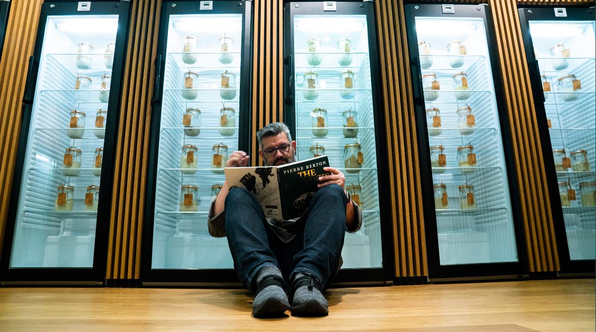 a man sits on the floor, reading a book in front of glass door refrigerators that contain jars.