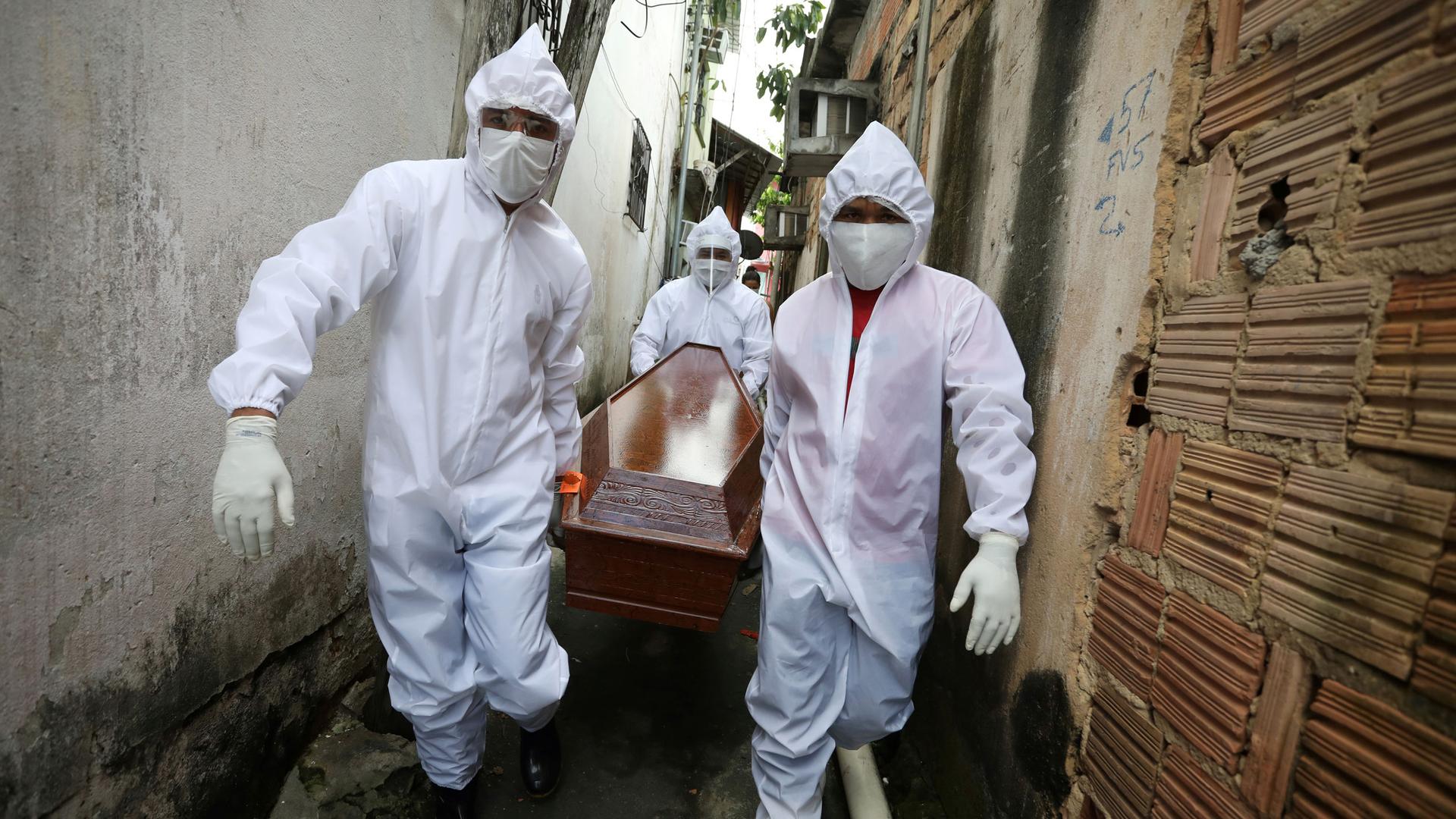 Three workers are shown wearing white medical protective clothing and masks while carrying a wooden coffing in a small alley. 