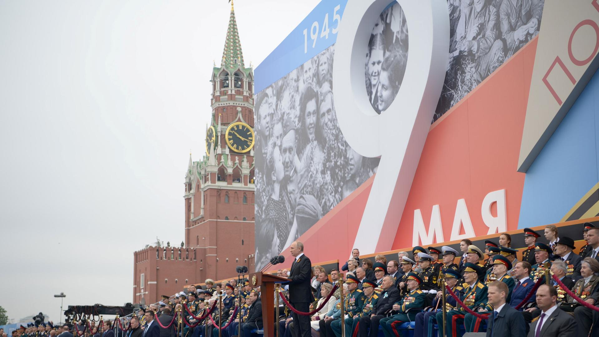 Putin stands at a podium in front of a crowd and signage for May 9