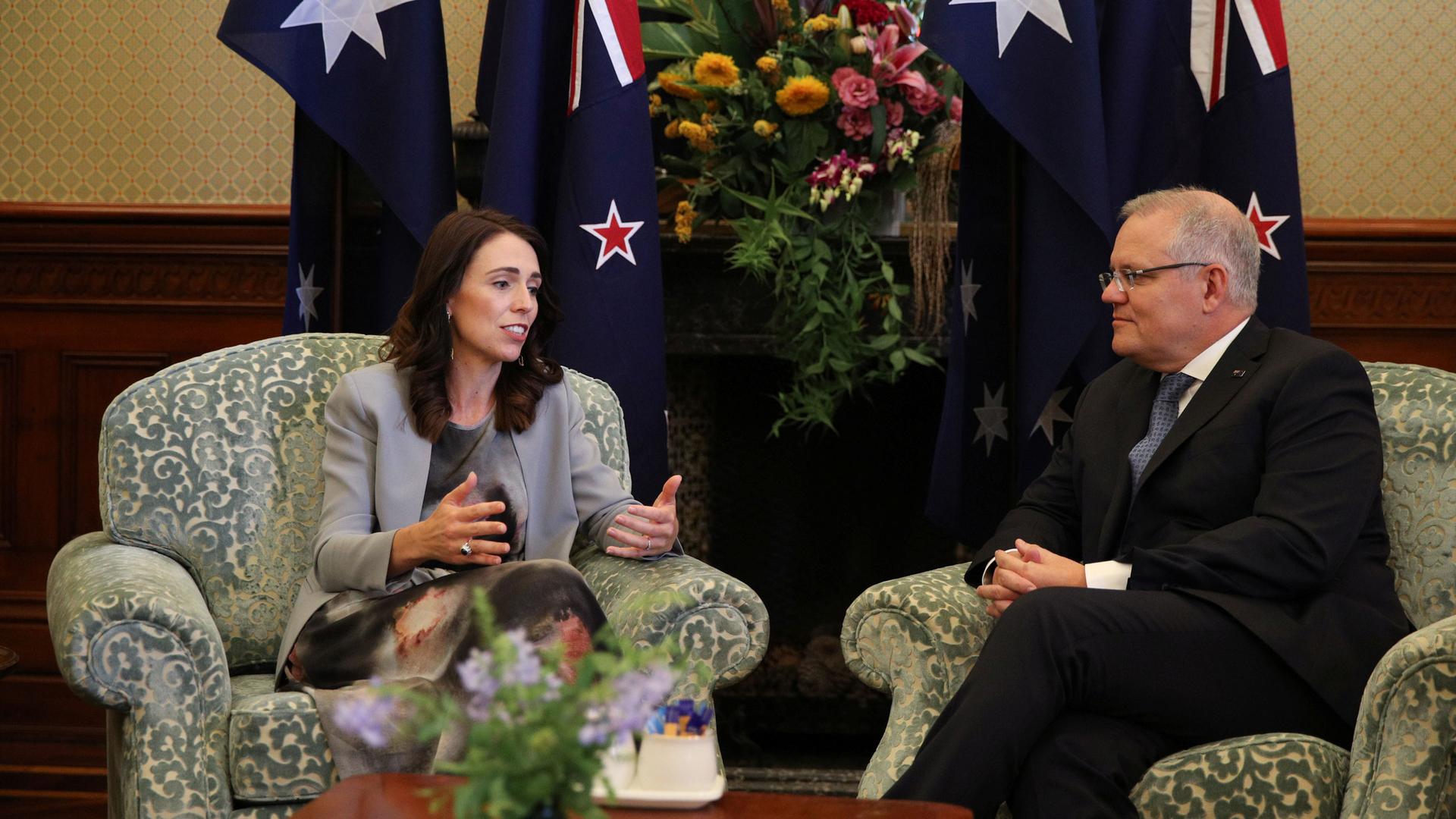 A woman and a man sit in chairs in front of the New Zealand and Australian flags