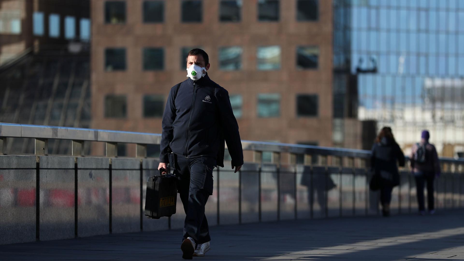 A man is shown wearing a white protective facemask and carrying a briefcase while walking.