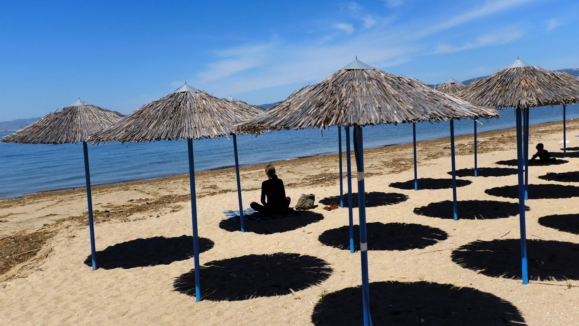 Several rows of thatch umbrellas are shown on a sandy beach with a woman in shadow sitting underneath one of the umbrellas.