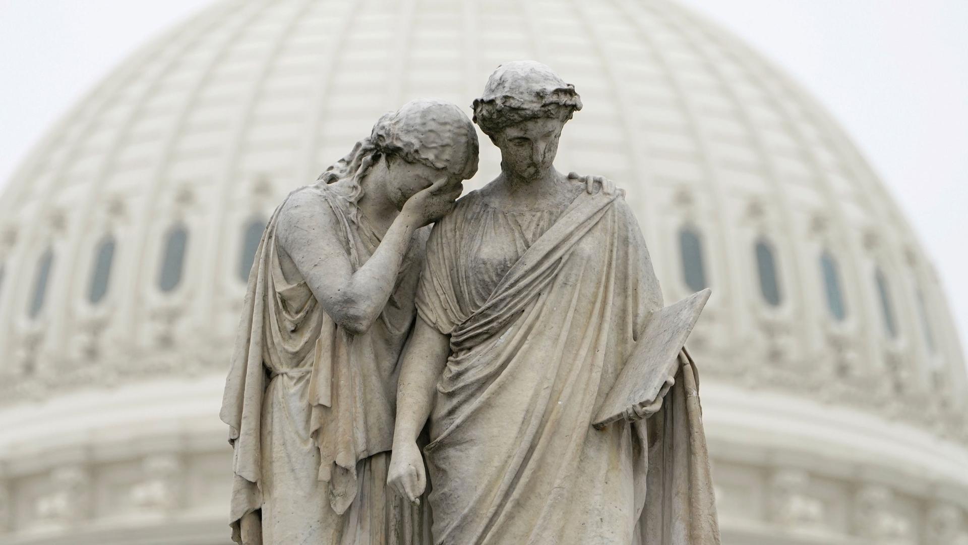 A statue is shown depicting two women with one holding her hand to her face and the other holding a book.