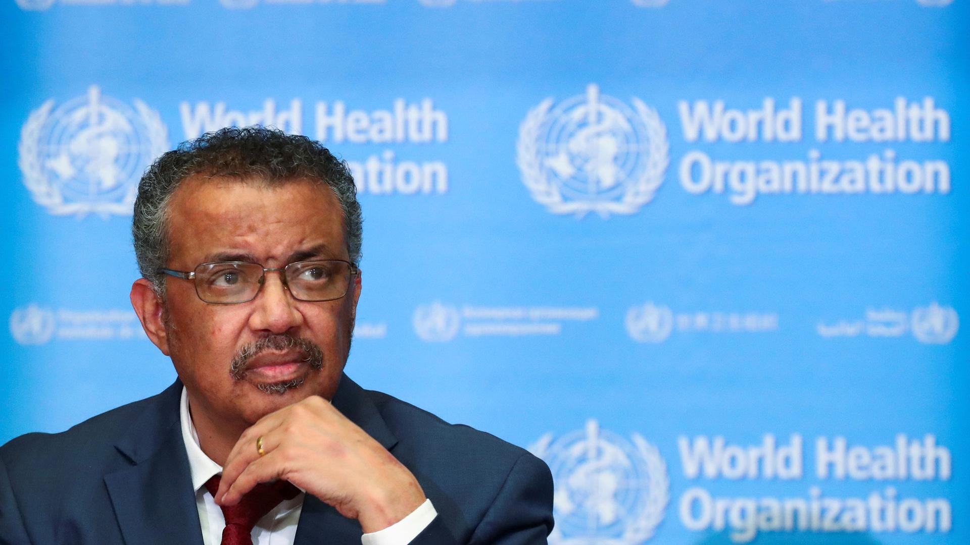 Tedros Adhanom Ghebreyesus is shown sitting with a red tied and jacket with his hand on his chin.