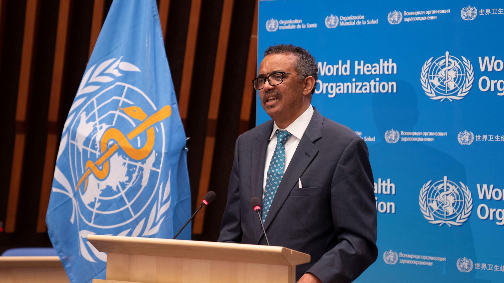 Tedros Adhanom Ghebreyesus is shown standing at a wooden podium with the blue WHO flag next to him.