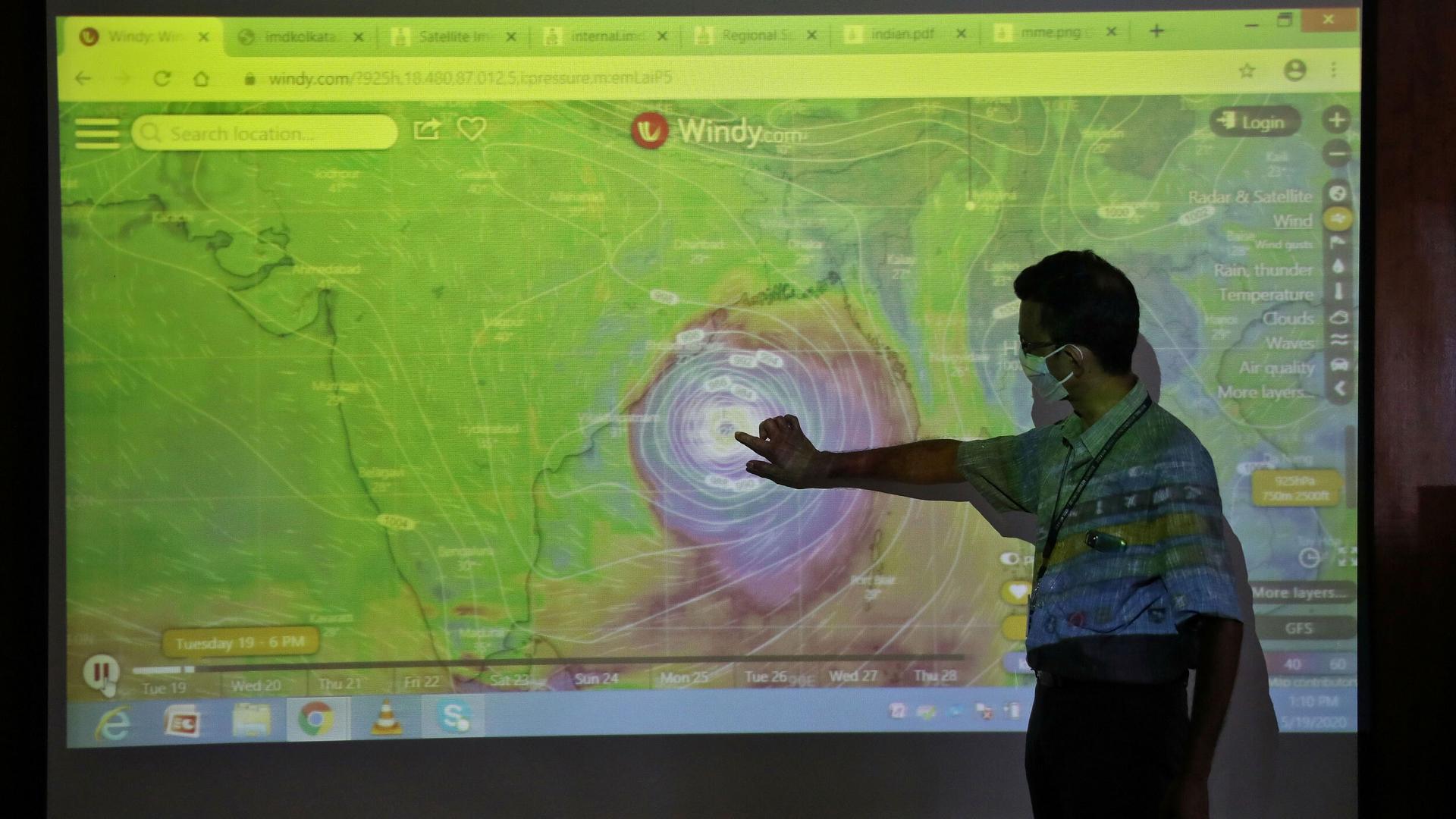 A man is shown wearing a protective face mask and pointing to an image of a cyclone projected on a screen.