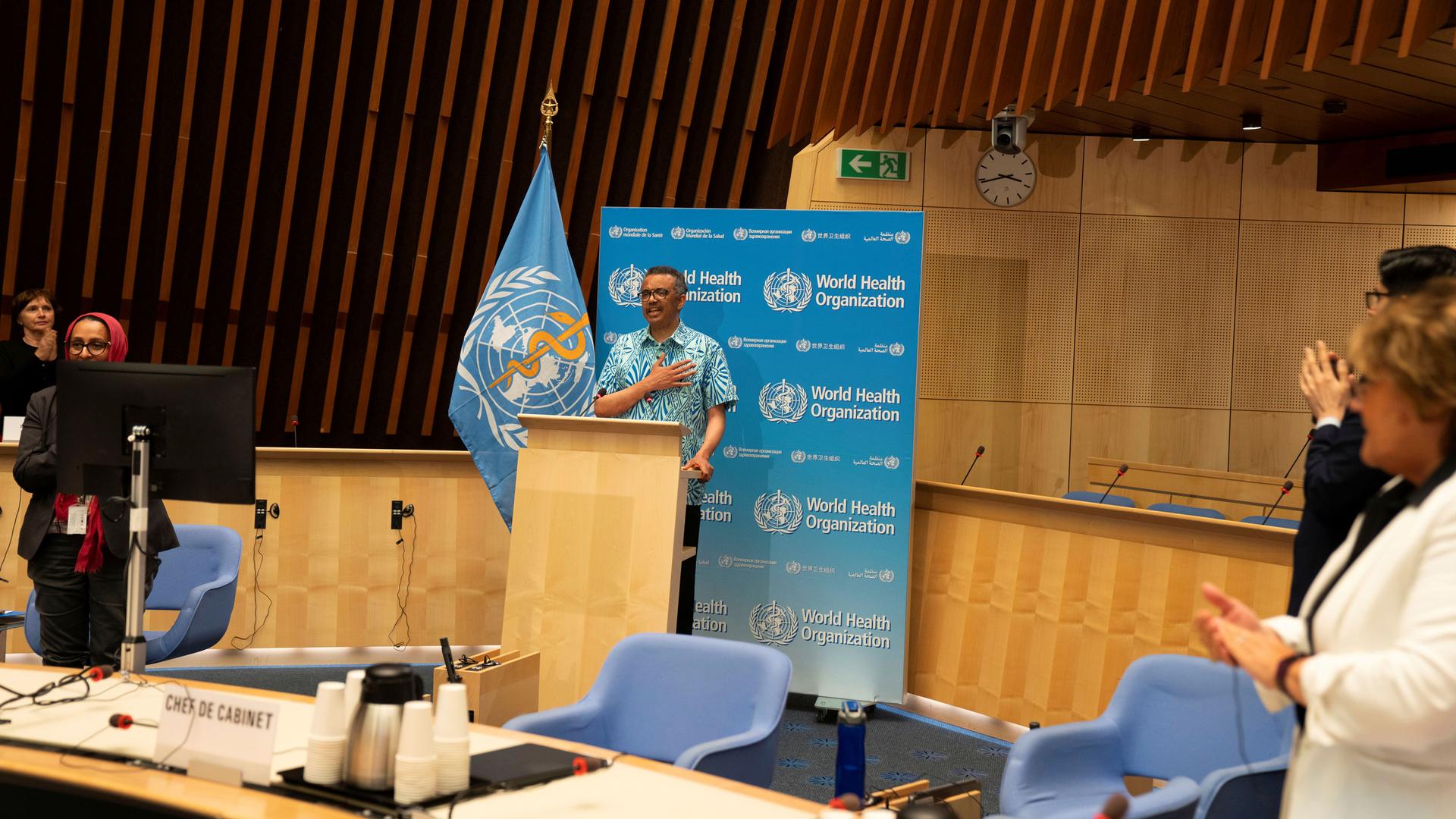Tedros Adhanom Ghebreyesus is shown standing at a wooden podium with the blue WHO flag next to him and wearing a blue-print button down short-sleeved shirt.