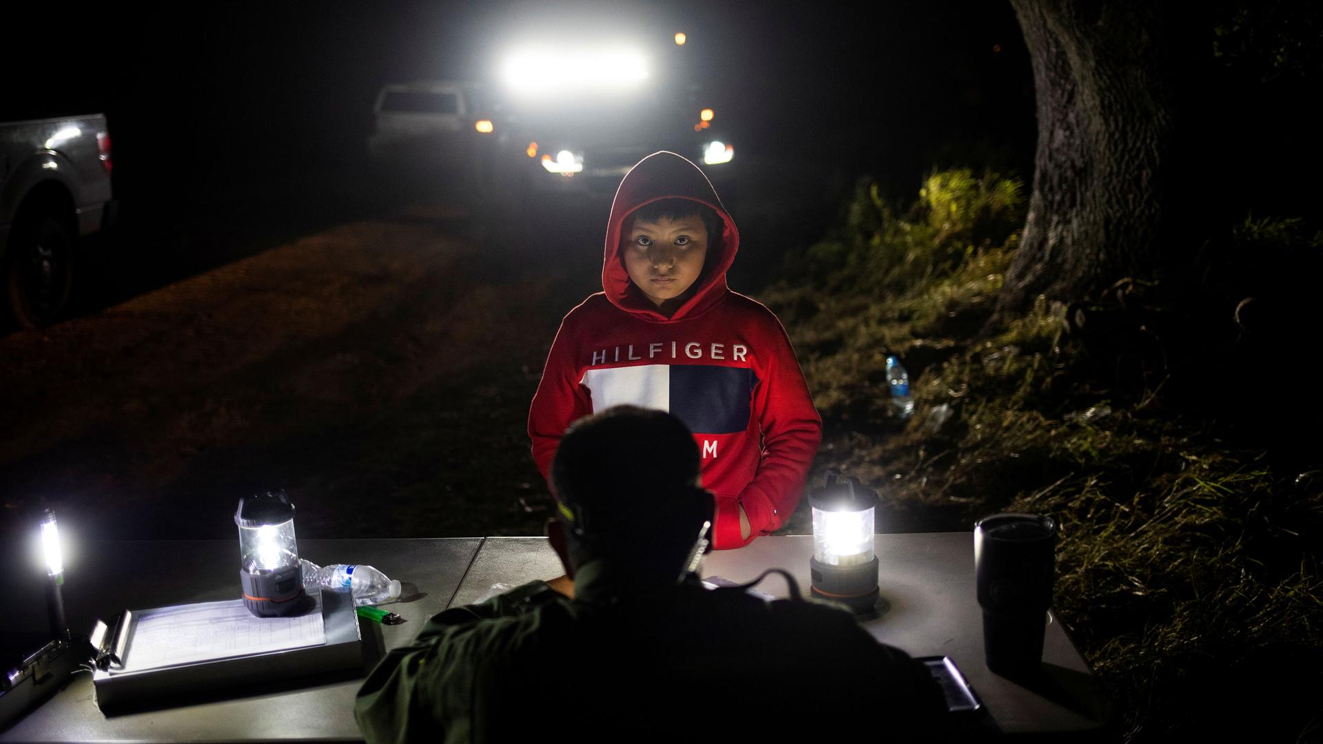 A young boy is shown wearing a red hooded sweatshirt and standing at a border patrol table.