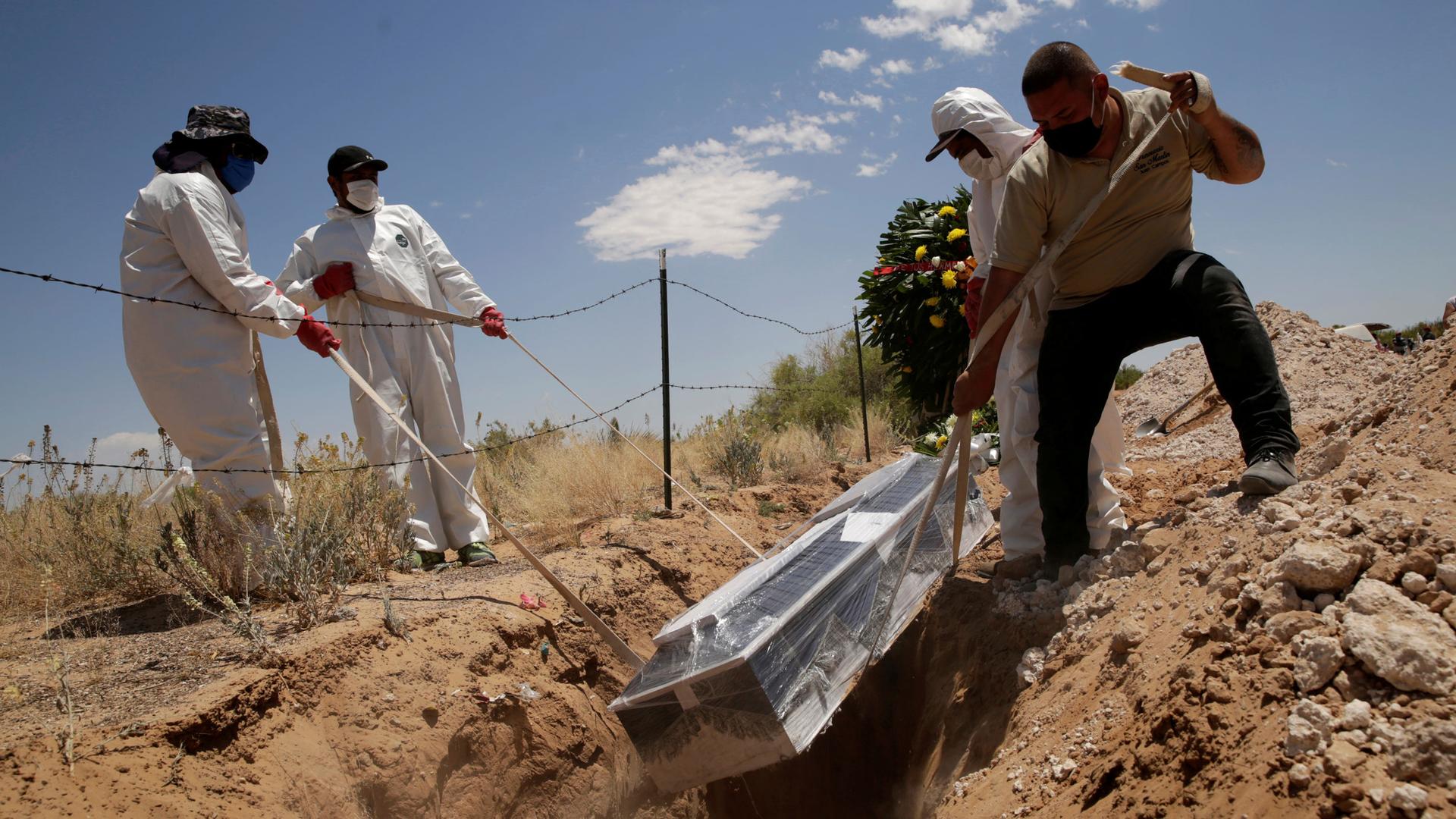 For men are shown wearing protective clothing and lowering a casket wrapped in clear plastic into a grave.