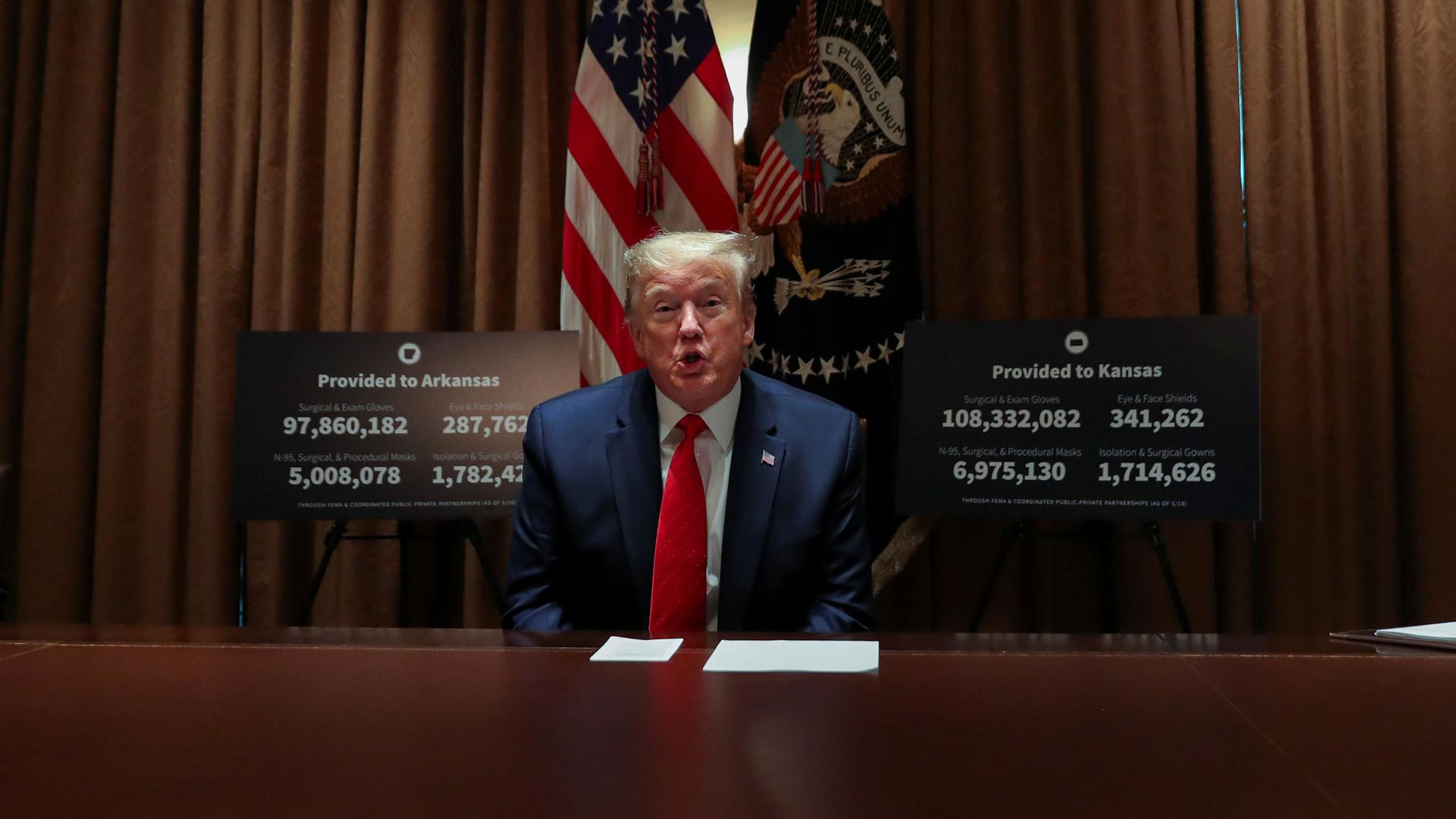 US President Donald Trump is shown sitting at a table wearing a dark suit and red tied with the US flag behind him.