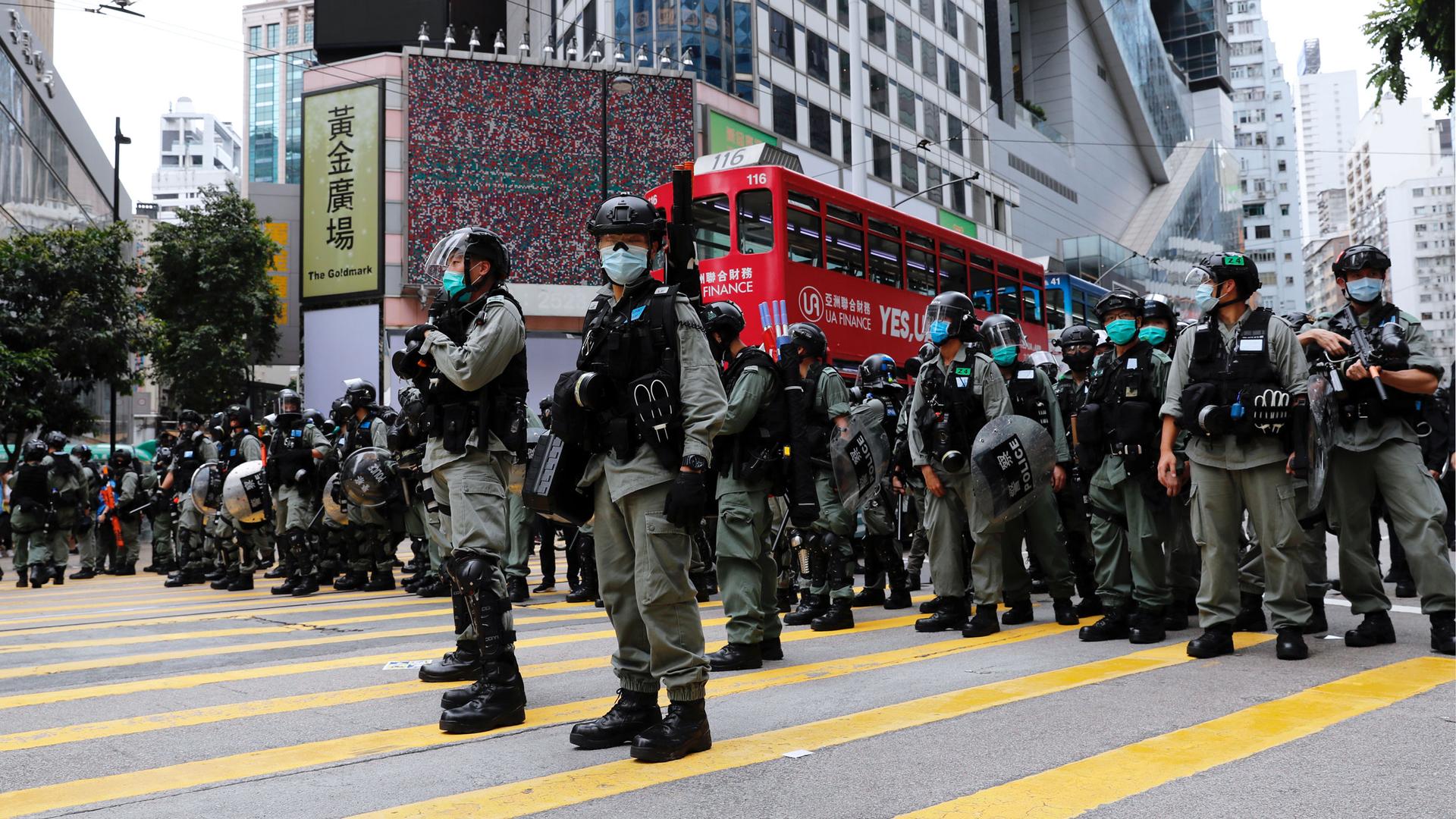 Riot police are shown standing in a line across the middle of a large street and wearing protective armor in Hong Kong.