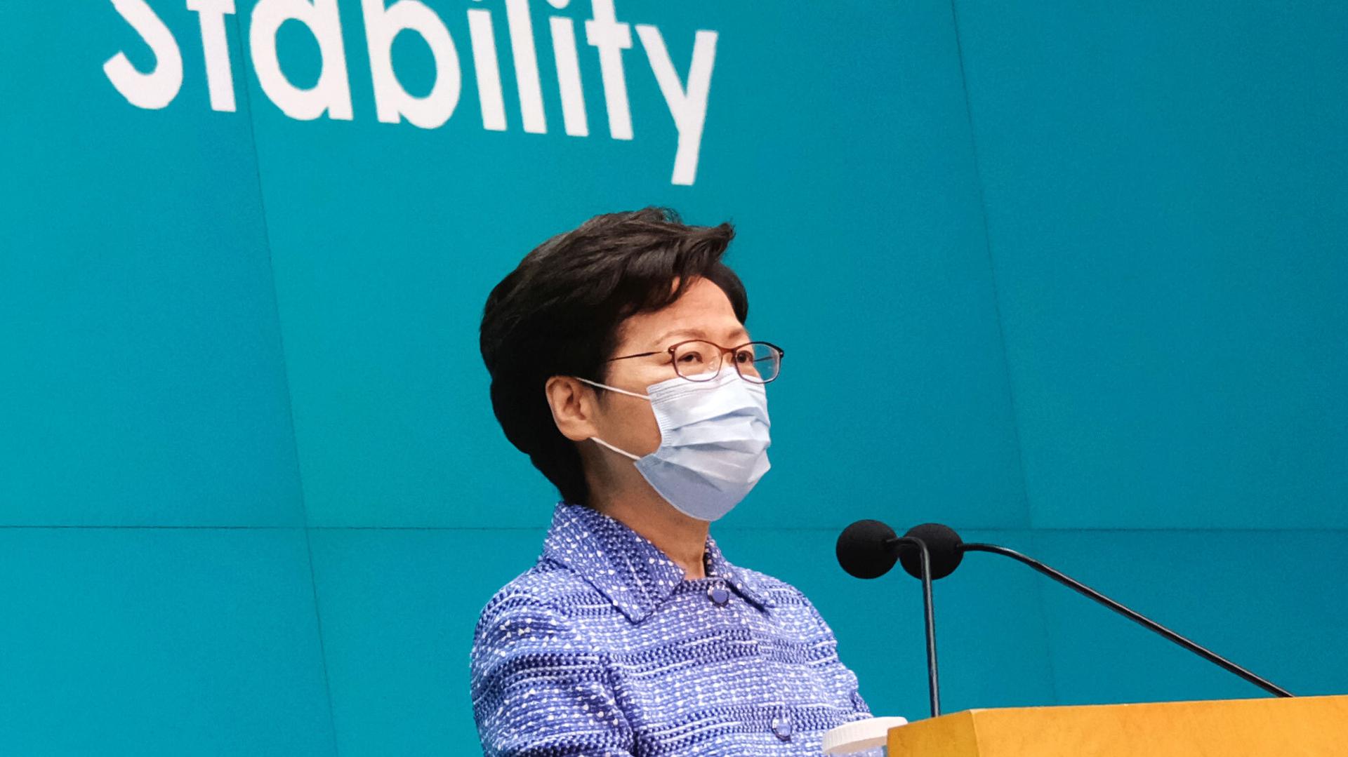 Hong Kong Chief Executive Carrie Lam is shown standing at a wooden podium with two small microphones and wearing a protective face mask.