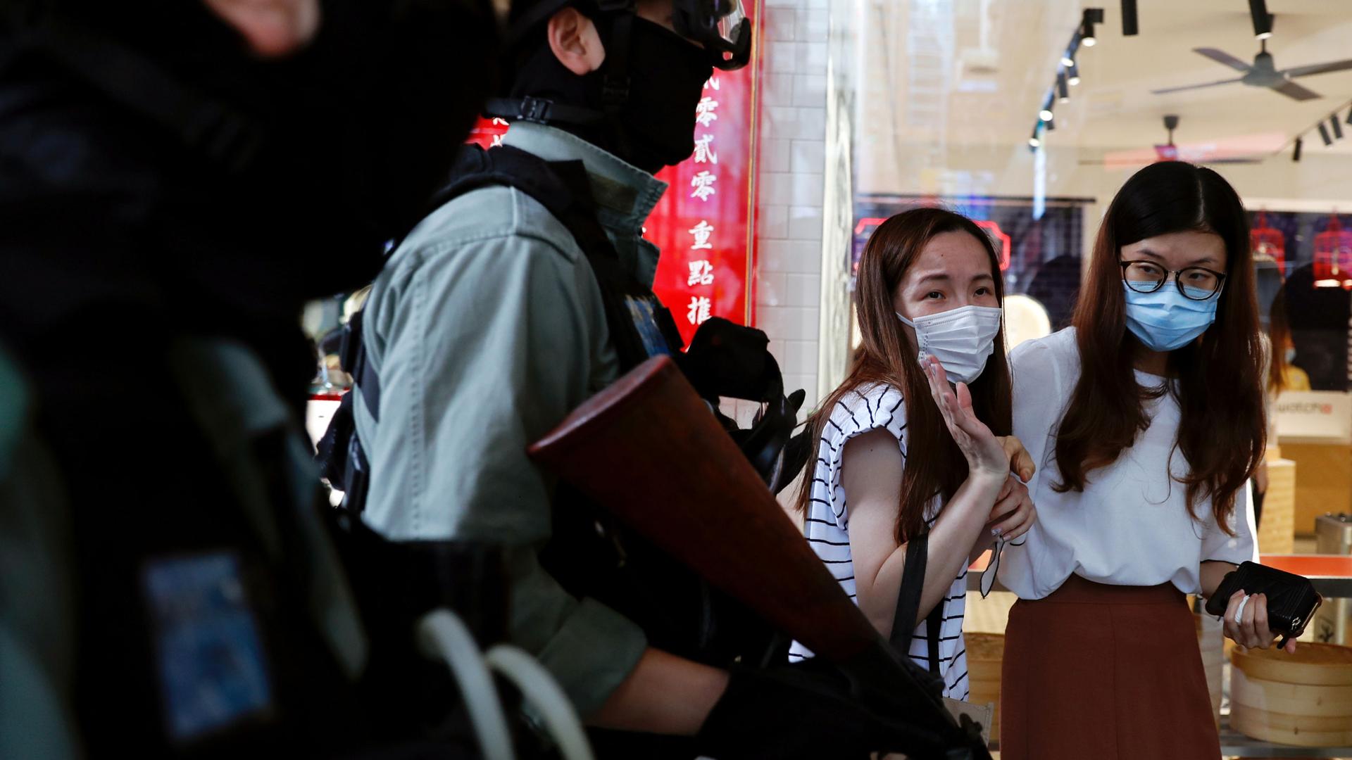 Two women are shown wearing protective face masks as several police officers walk past carrying weapons and wearing protective riot gear.