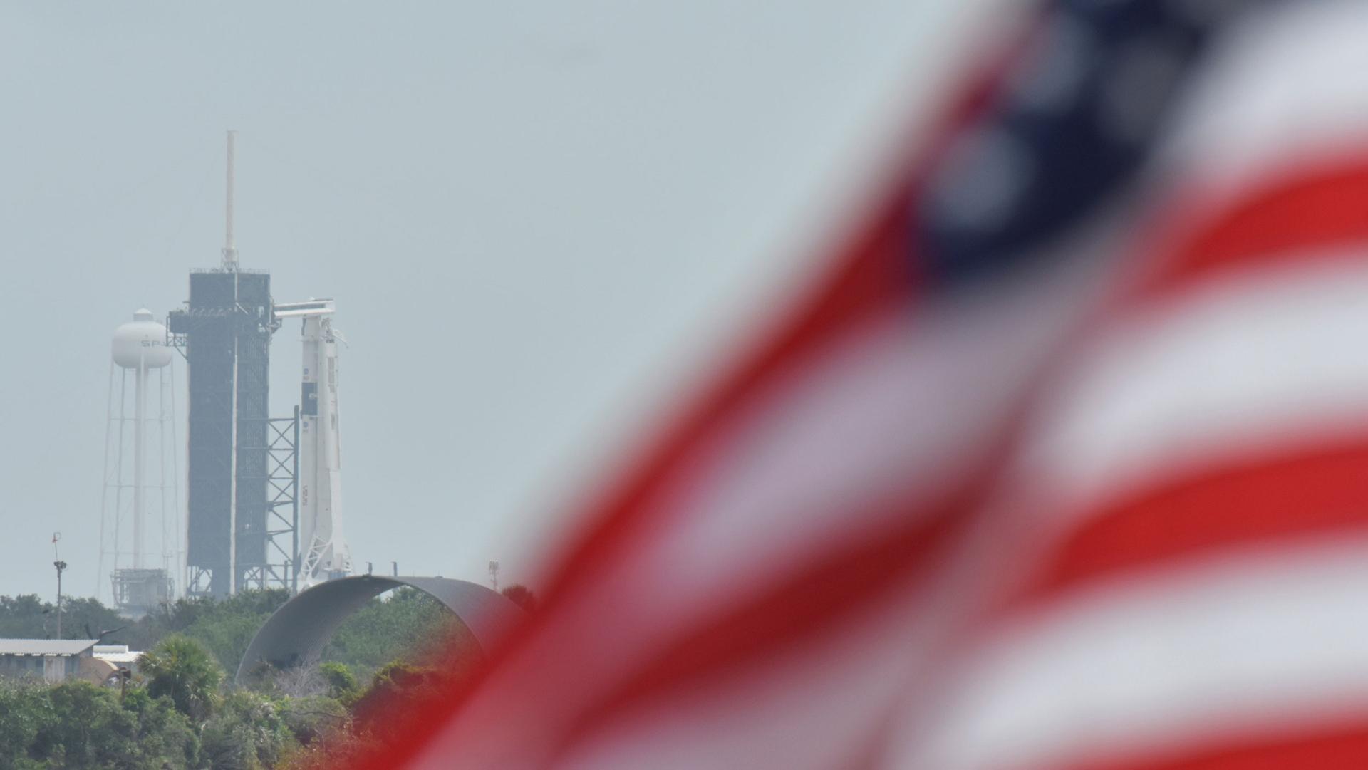 A US flag flutters in the breeze. In the background, a rocket launchpad