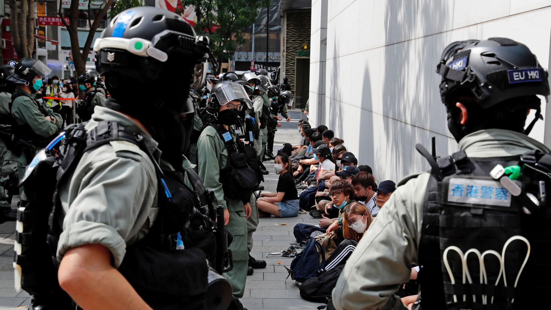 A line of young demonstrators are shown sitting on the ground next to a building with riot police standing in front of them.