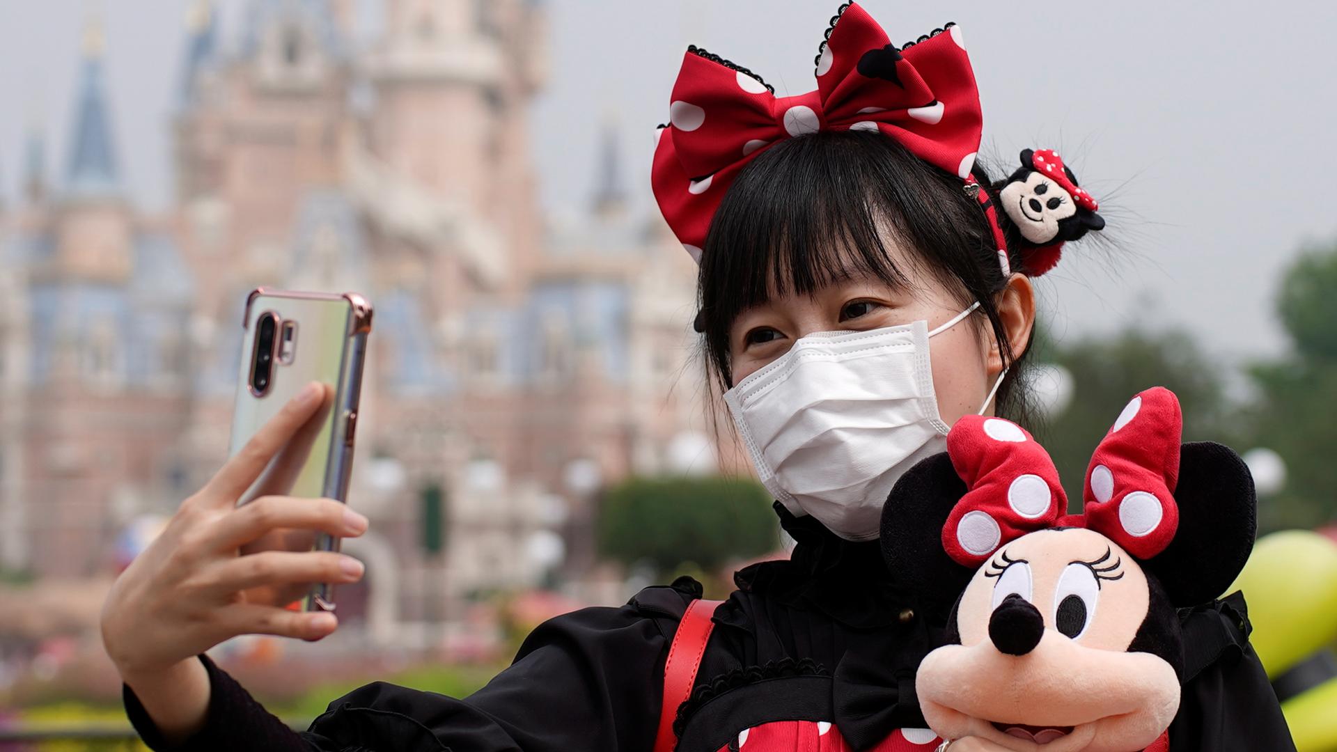 A young woman wears a red bow and holds a doll as she takes a selfie at Disneyland in Shanghai
