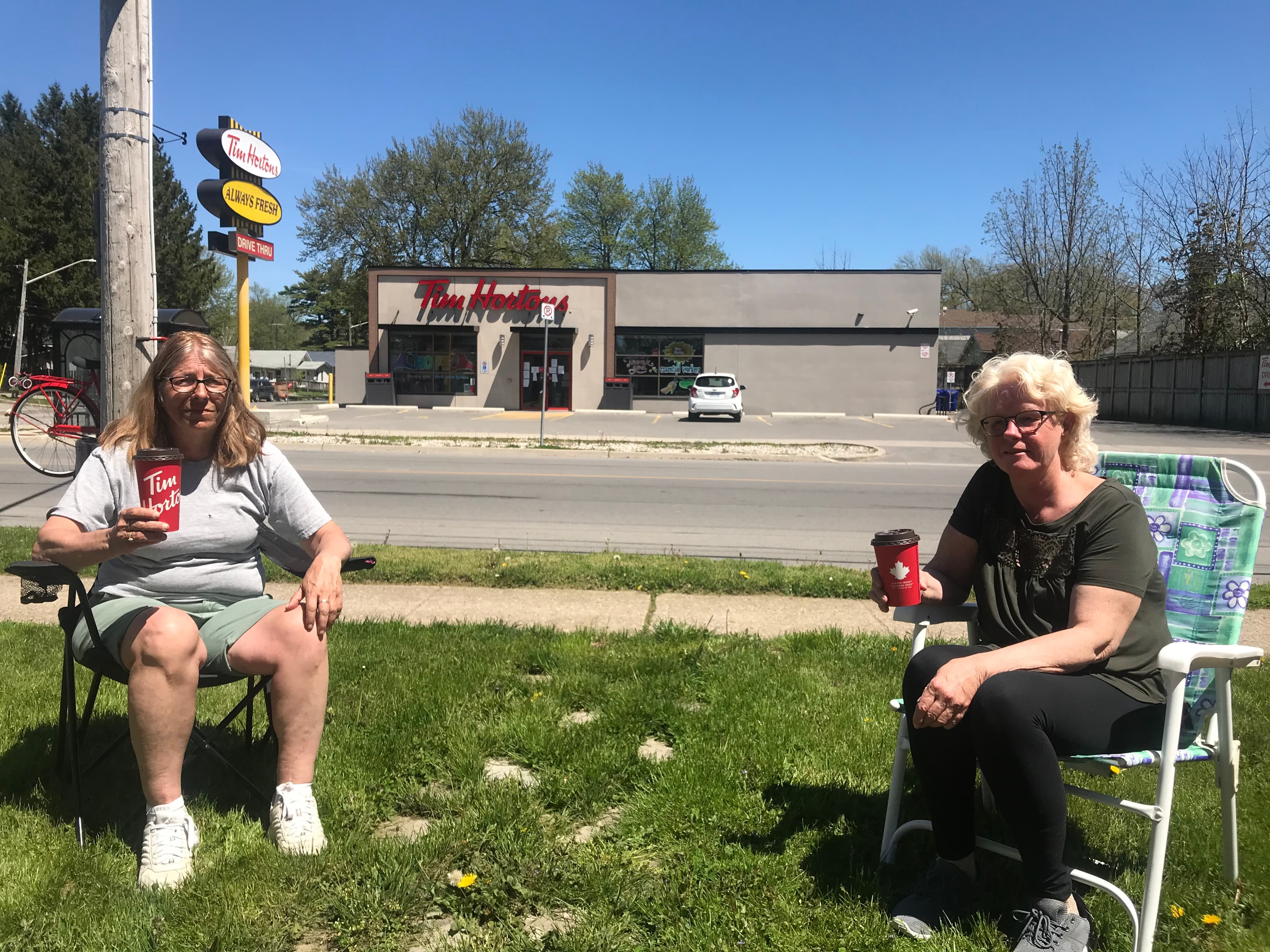 Two women sit in lawn chairs and drink a coffee near a coffee shop.