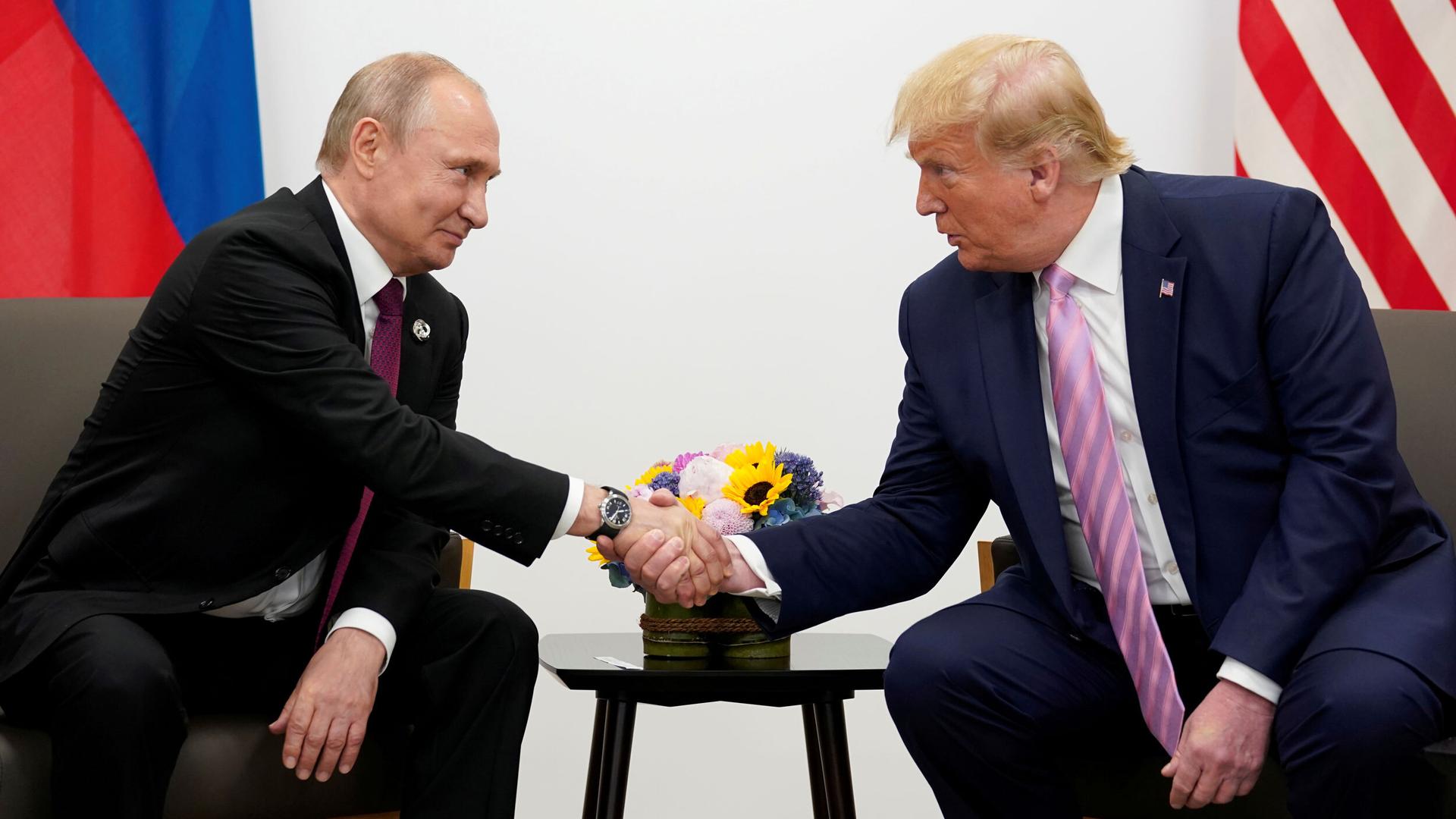 Russia’s President Vladimir Putin and US President Donald Trump are shown sitting in arm chairs and leaning toward each other shaking hands.
