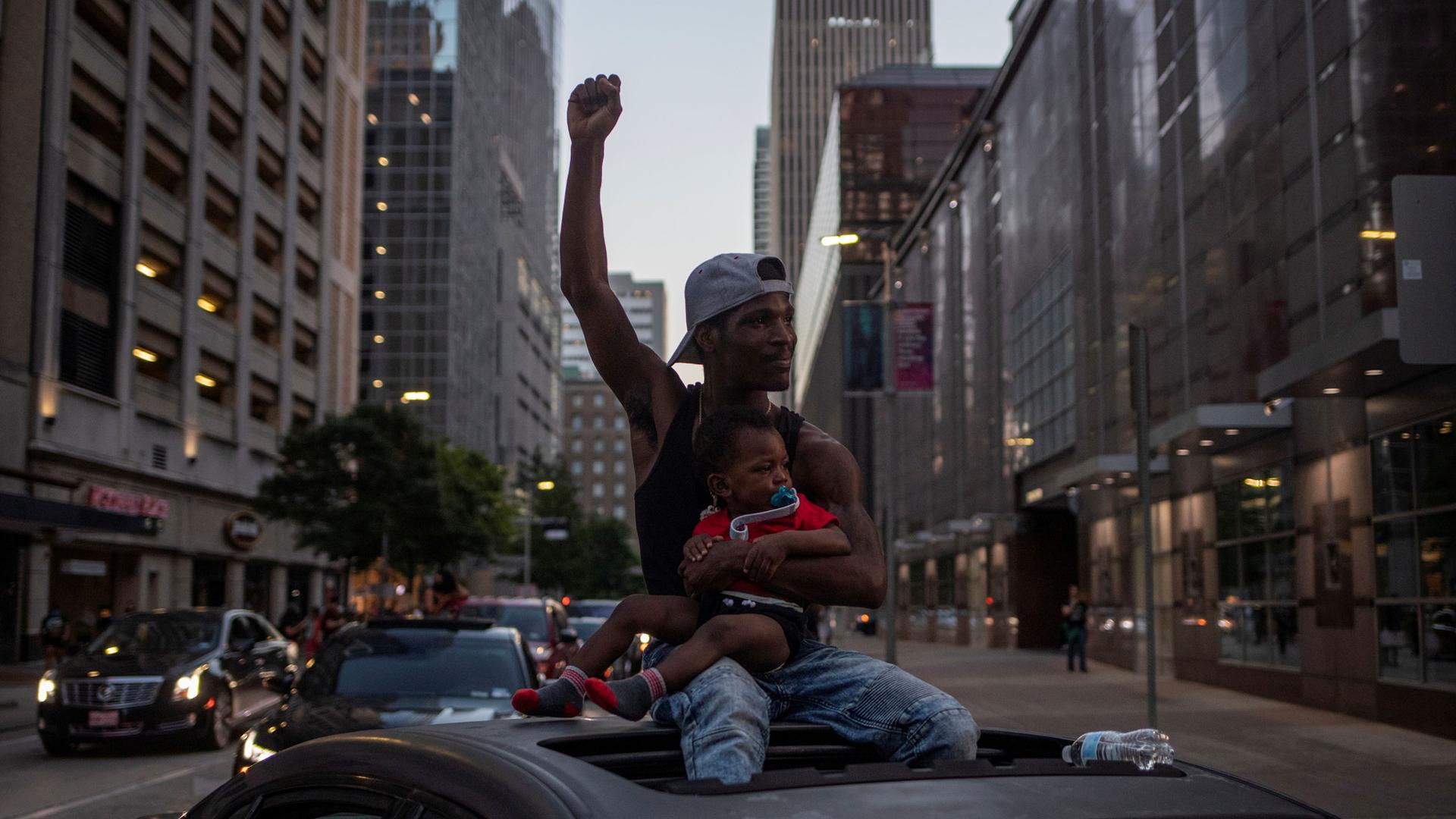 A man is shown sitting on the roof of his car through the sunroof and hold a young child in his lap while raising his fist.