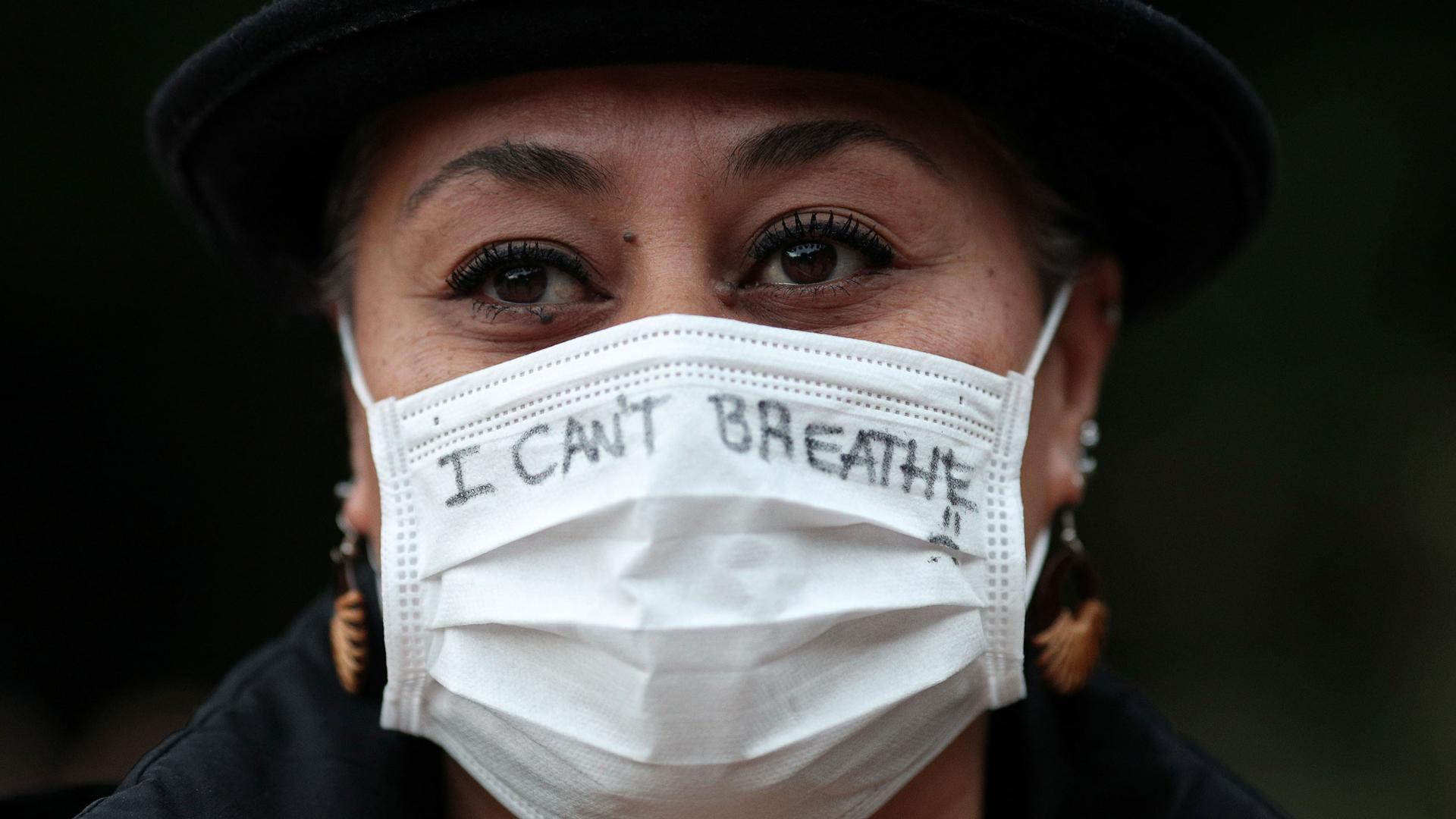 A close up photograph of a woman wearing a white protective face mask with the words, "I can't breathe" written on it.