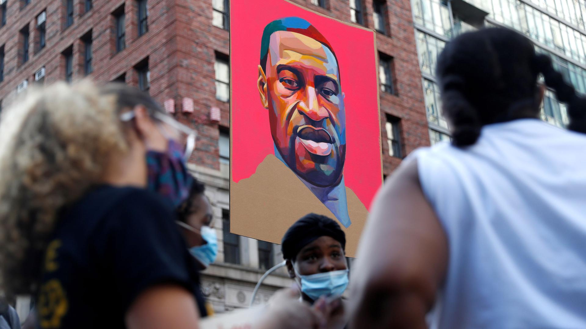 A portrait of George Floyd is seen hanging in the background as demonstrators in the near ground are in soft focus.