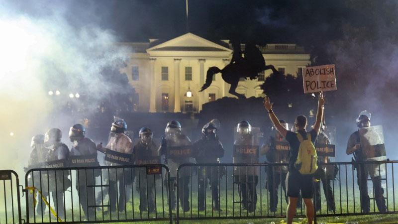 Smoke in front of the White House as riot police stand with a protester in the foreground.
