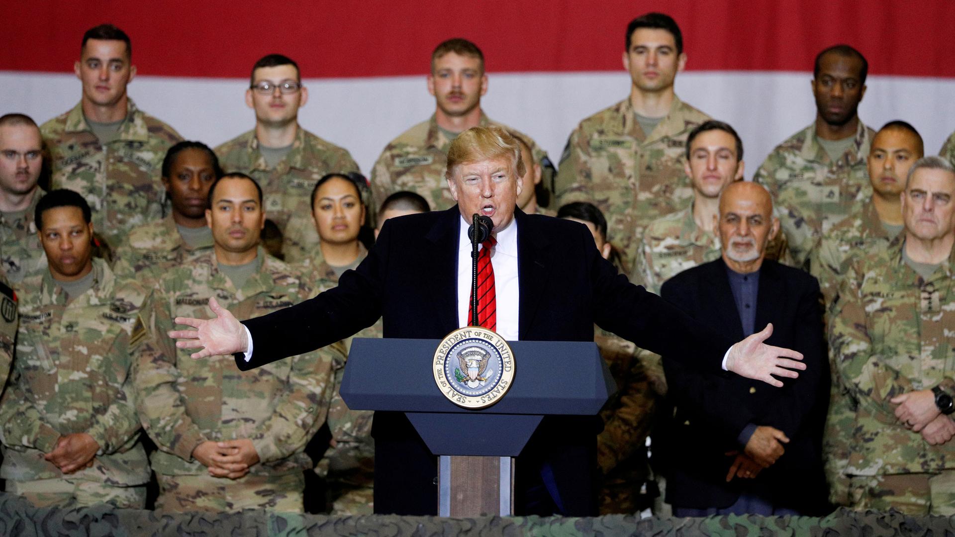 US President Donald Trump is shown standing at a podium with his arms outstretched and rows of US soldiers wearing fatigues standing behind him.