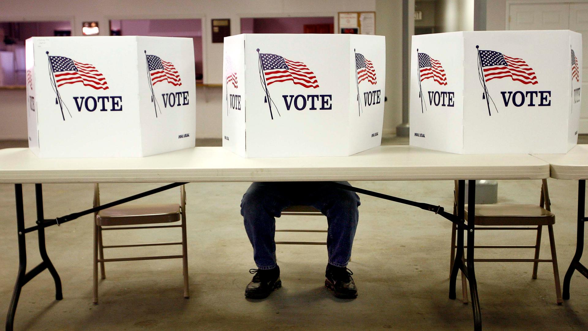 Una persona esta sentada dentro de una urna de votacion, cubierta con carteles que dicen "vote" y con banderas de los Estados Unidos.